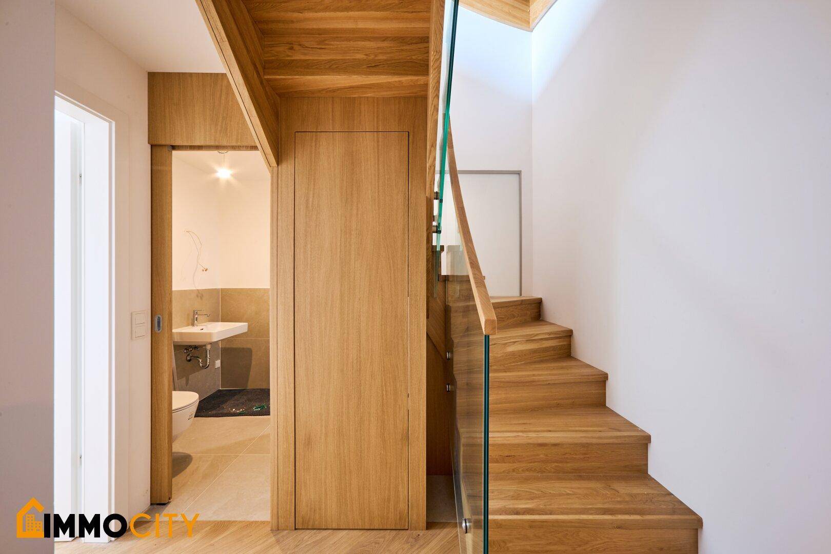 Helle Holztreppe mit elegantem Glasgeländer und angrenzendem, modern ausgestattetem Gäste-WC.