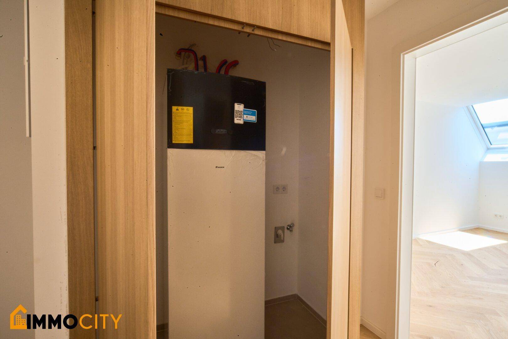 Versteckter Technikraum mit Warmwasserbereiter, integriert in eine Holzwand, neben einem hellen Raum mit Dachfenster.