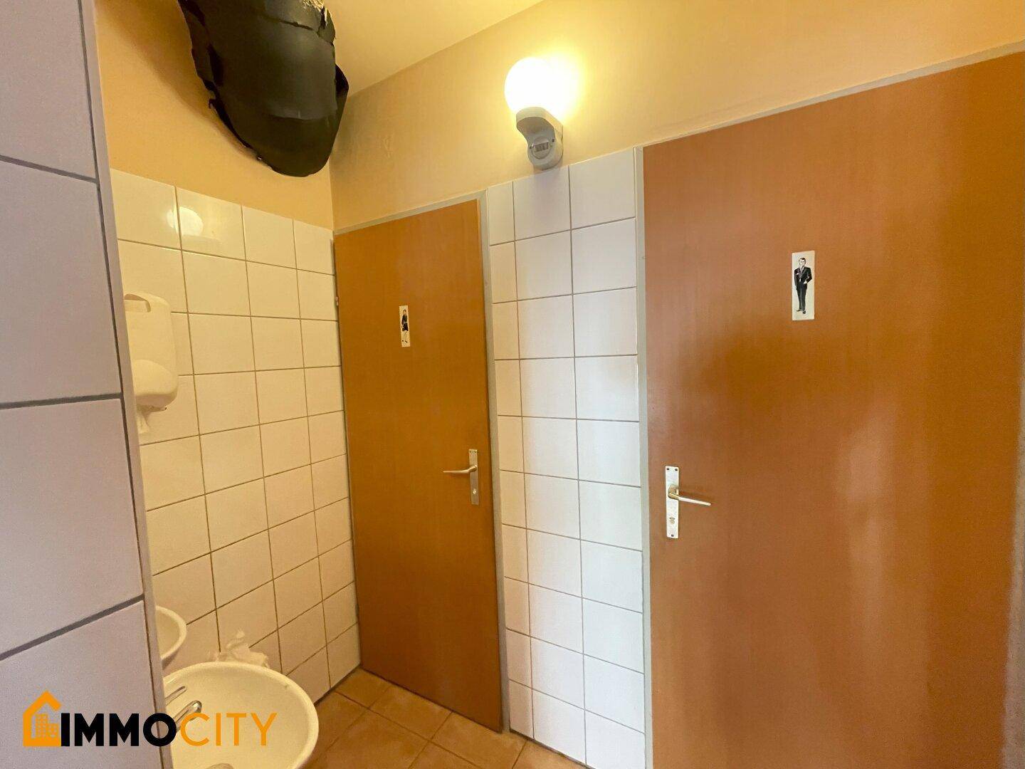 Öffentliche Toiletten mit zwei Kabinen, braunen Türen, weißen Wandfliesen und einem Waschbecken.