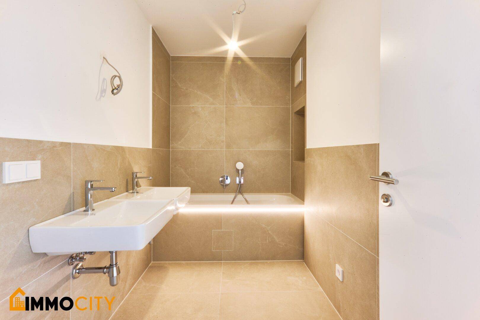 Modernes Badezimmer mit beigefarbenen Fliesen, Doppelwaschbecken und beleuchteter Badewanne.