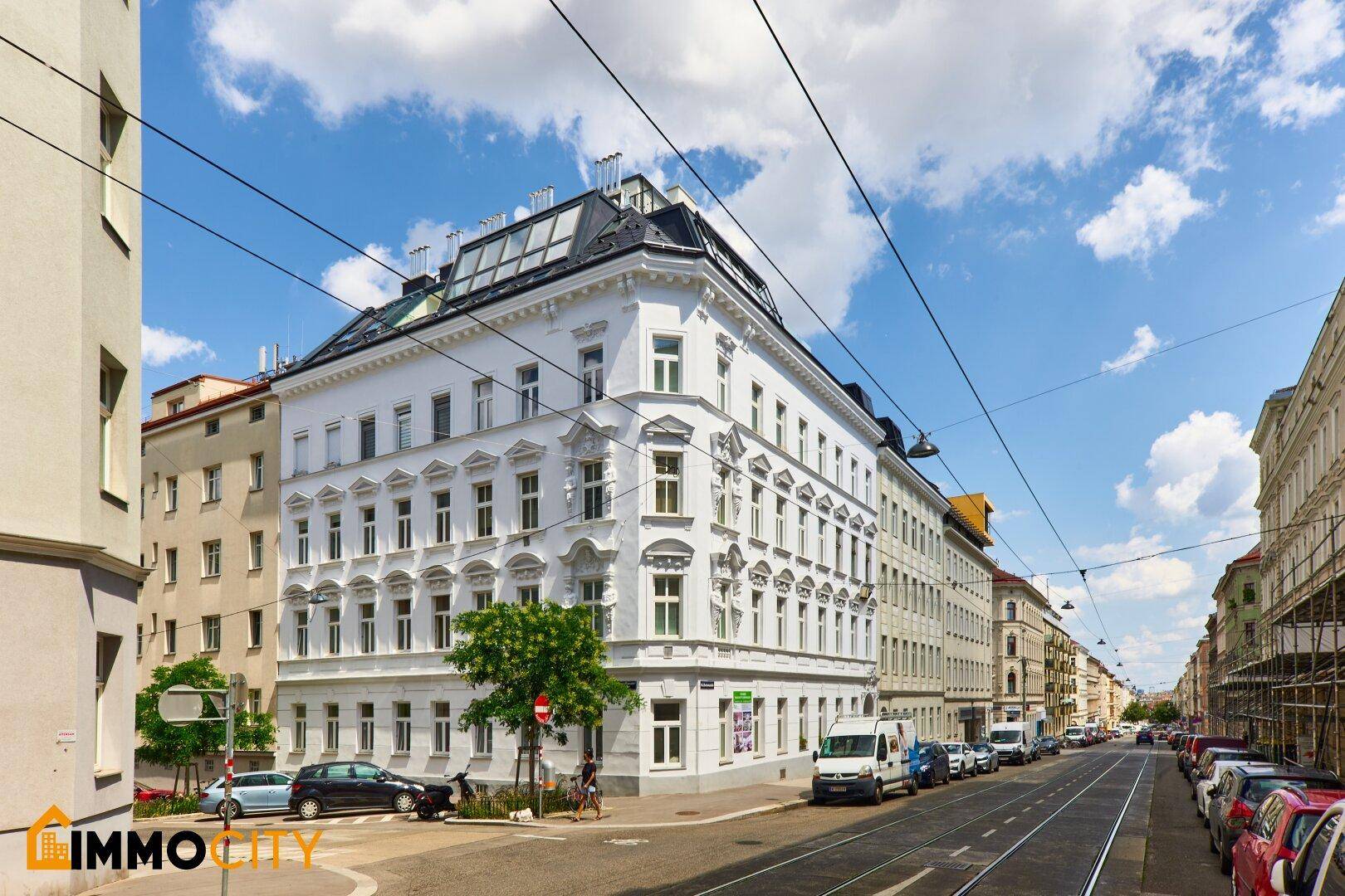 Weißes Eckgebäude mit klassischer Architektur und Mansarddach an einer belebten Straße.