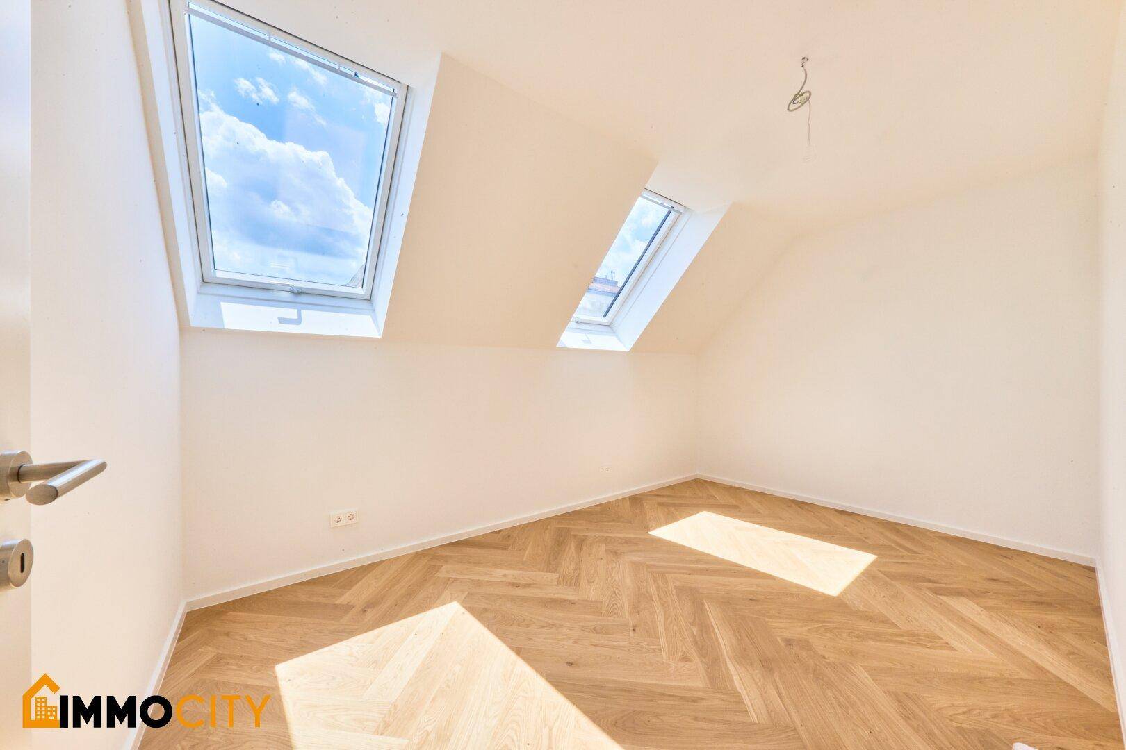 Heller Raum mit Dachschrägen, Fischgrätparkett und zwei Dachfenstern für natürliches Licht.