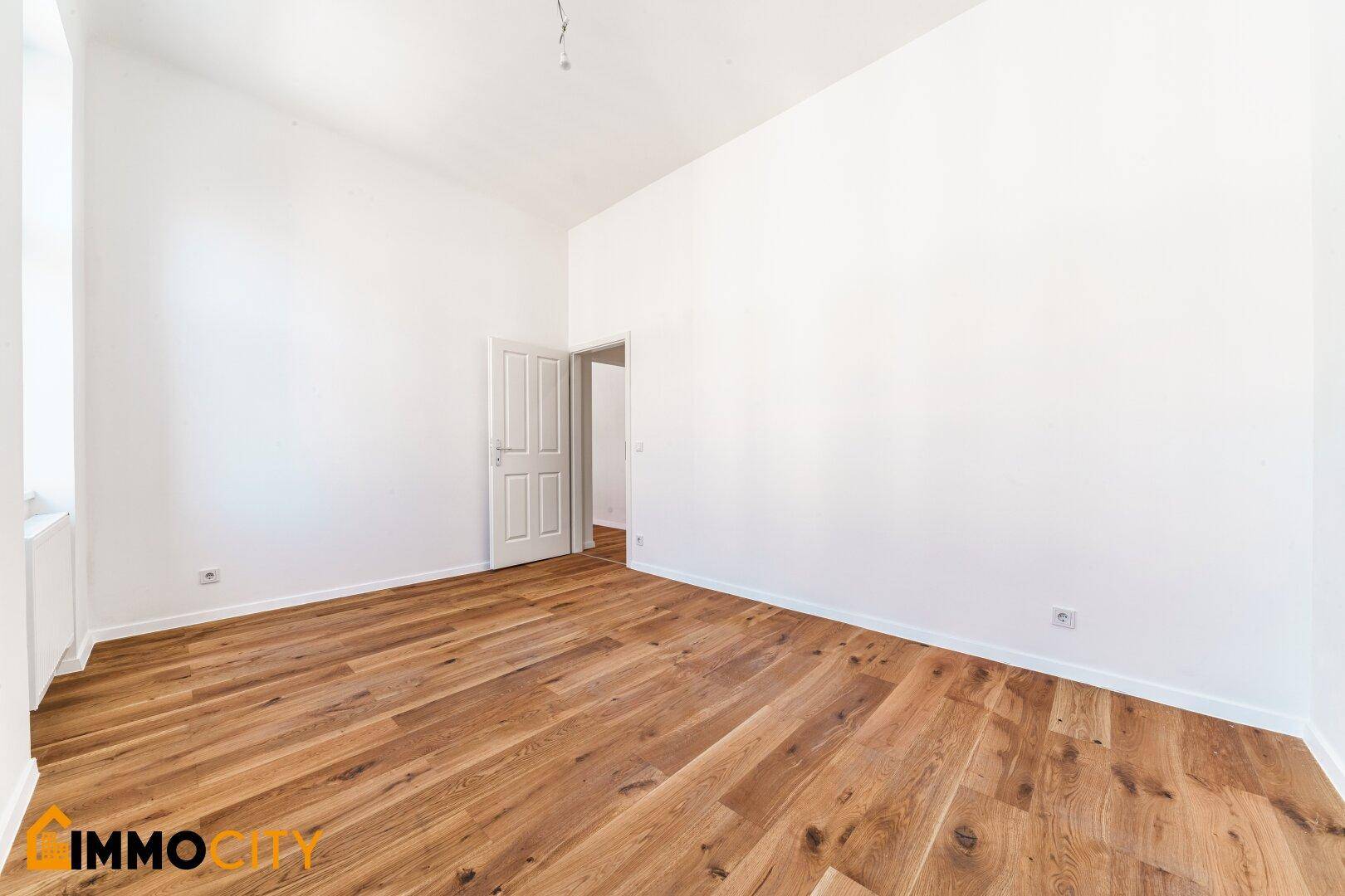 Helles Zimmer mit Parkettboden und Zugang zu weiteren Räumen, frisch gestrichen.