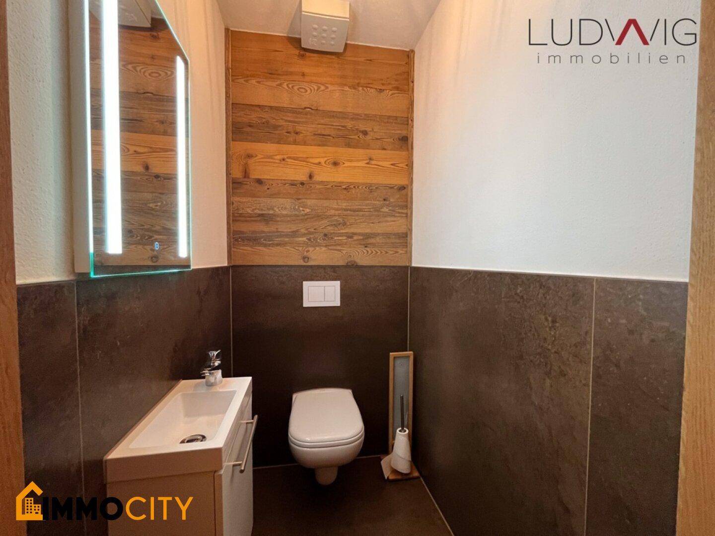 Modernes Gäste-WC mit Wand-WC, kleinem Waschbecken und beleuchtetem Spiegel.