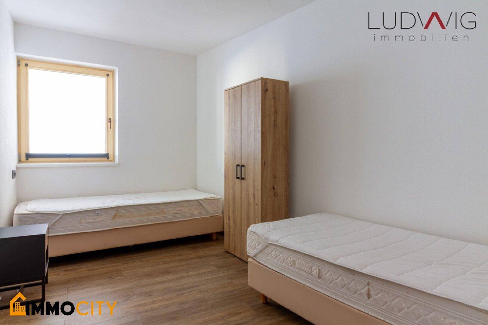 Helles Schlafzimmer mit zwei Einzelbetten, Holzschrank und Fenster mit Tageslicht.