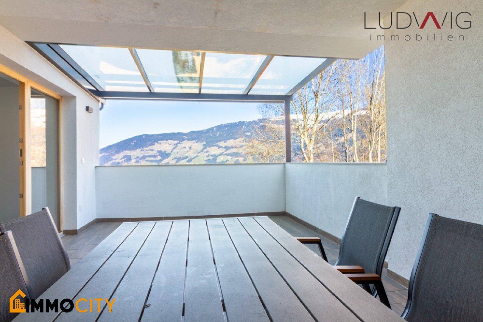 Überdachte Terrasse mit Holztisch und Stühlen, bietet Schutz und Bergblick.