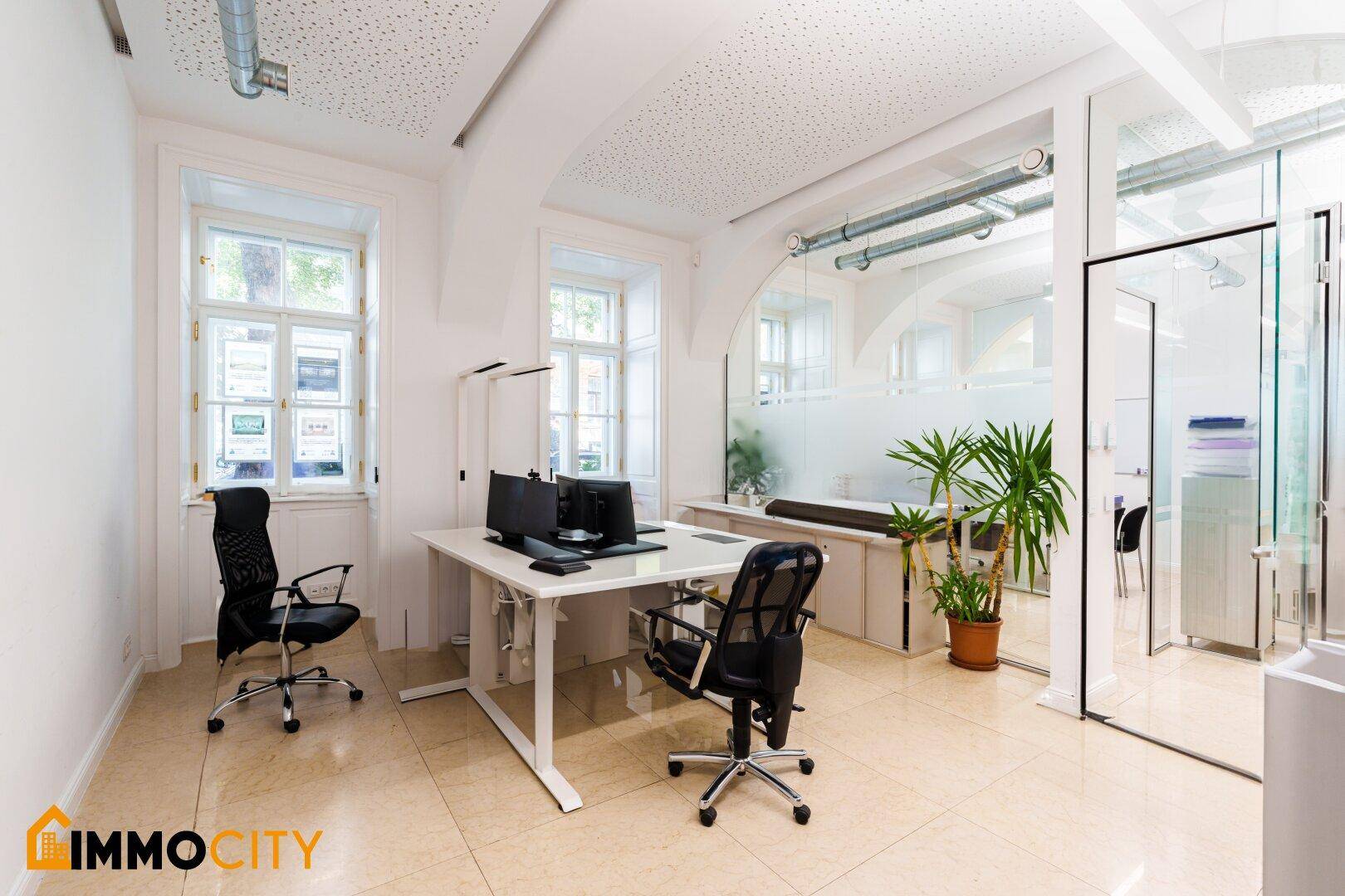 Moderner Büroraum mit Glaswänden, zwei Arbeitsplätzen und Pflanzen für eine angenehme Atmosphäre.