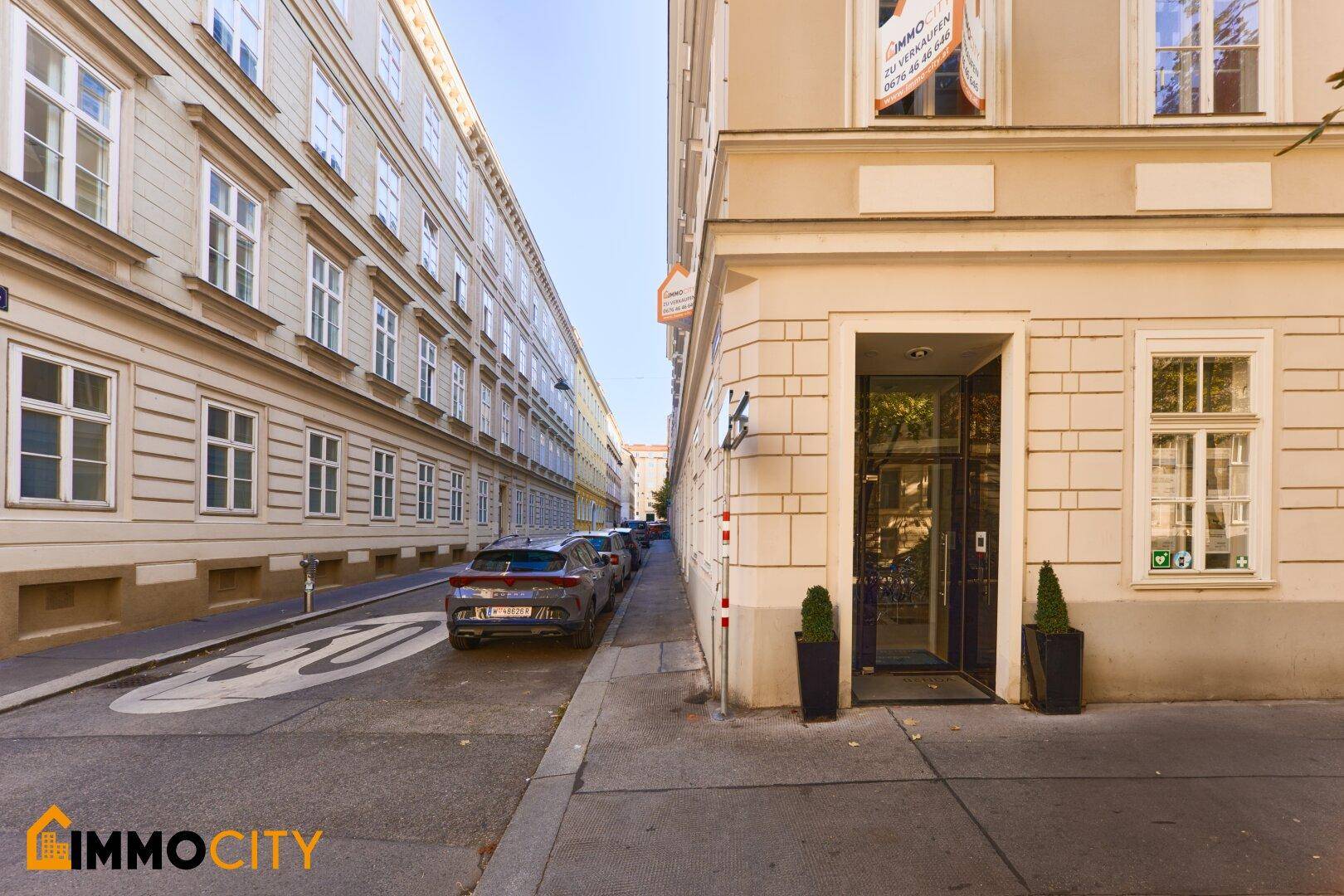 Historisches Gebäude an einer sonnigen Straße mit parkenden Autos und Bäumen.