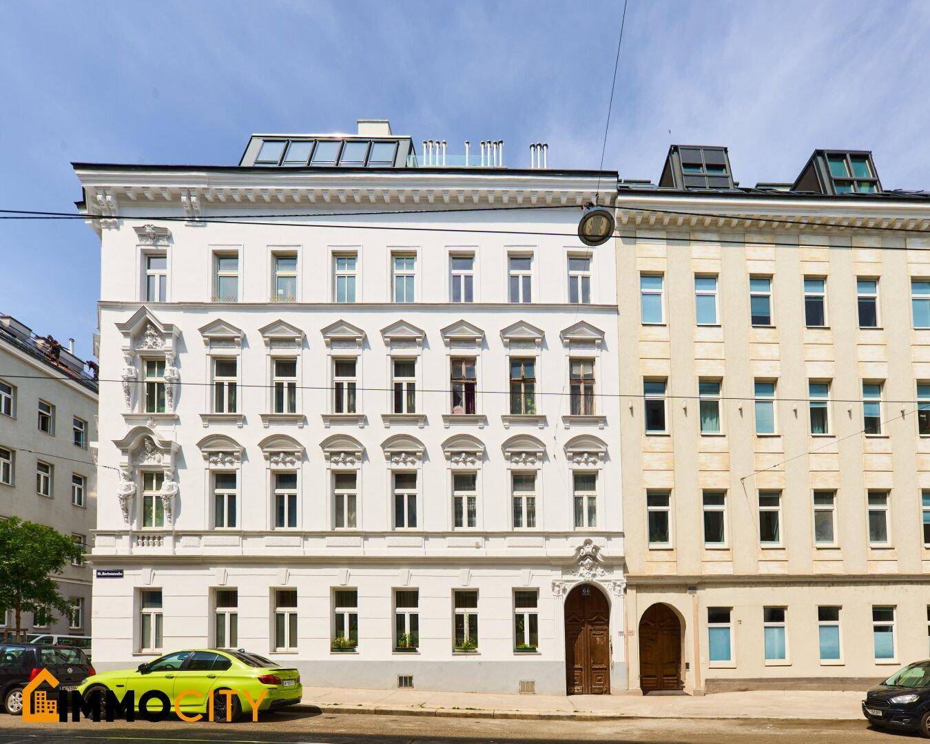 Historisches Gebäude mit weißer, verzierter Fassade und moderner Dachaufstockung in städtischer Umgebung.