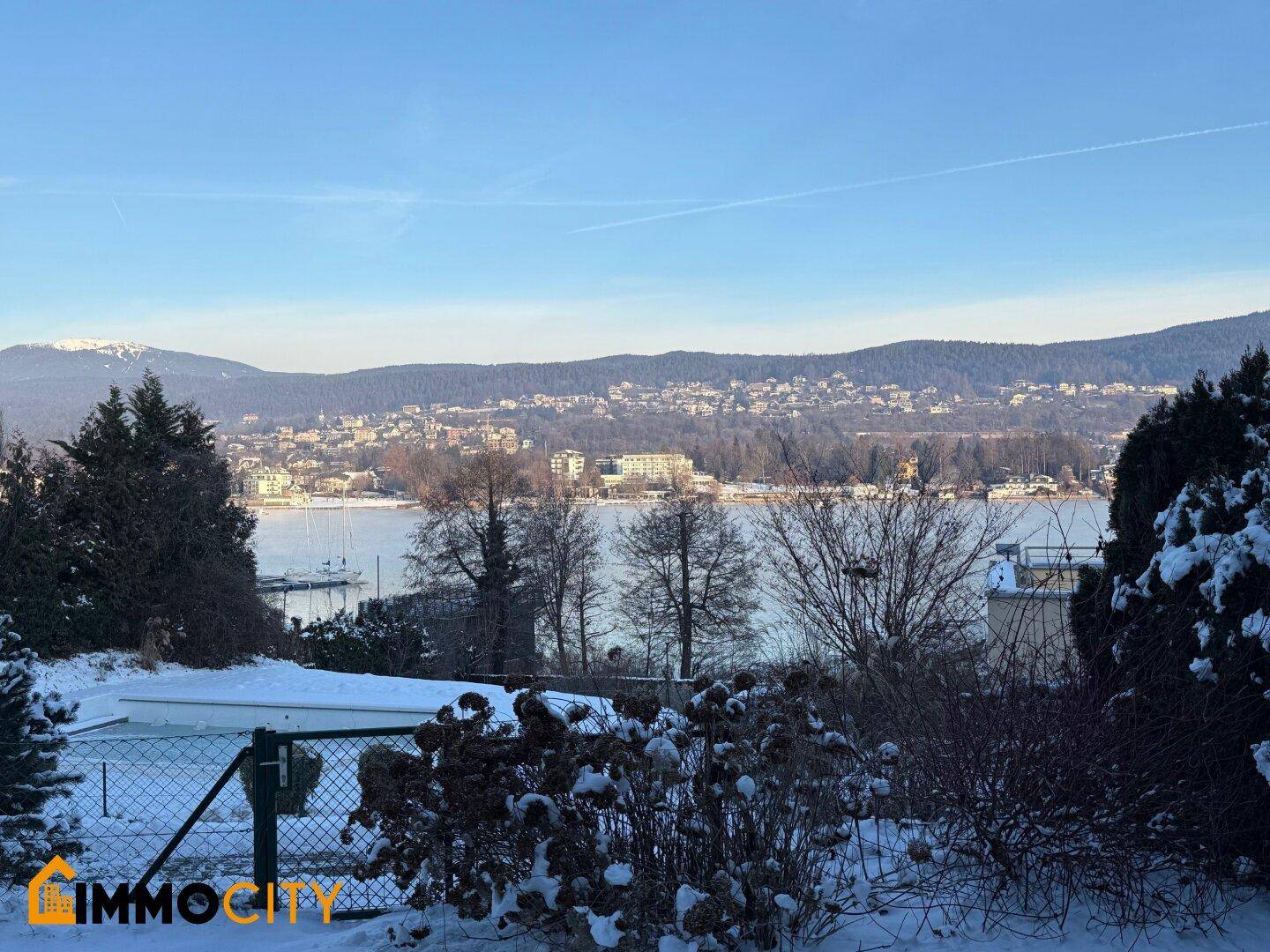 Panoramablick über den verschneiten Garten zum See und den bewaldeten Hügeln unter klarem Winterhimmel.