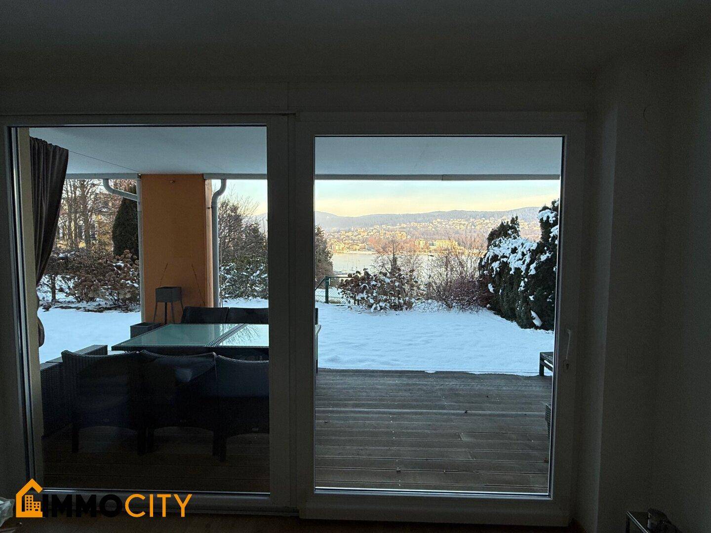 Blick aus dem Innenraum durch große Schiebetüren auf die Terrasse und den schneebedeckten Garten mit Seeblick.
