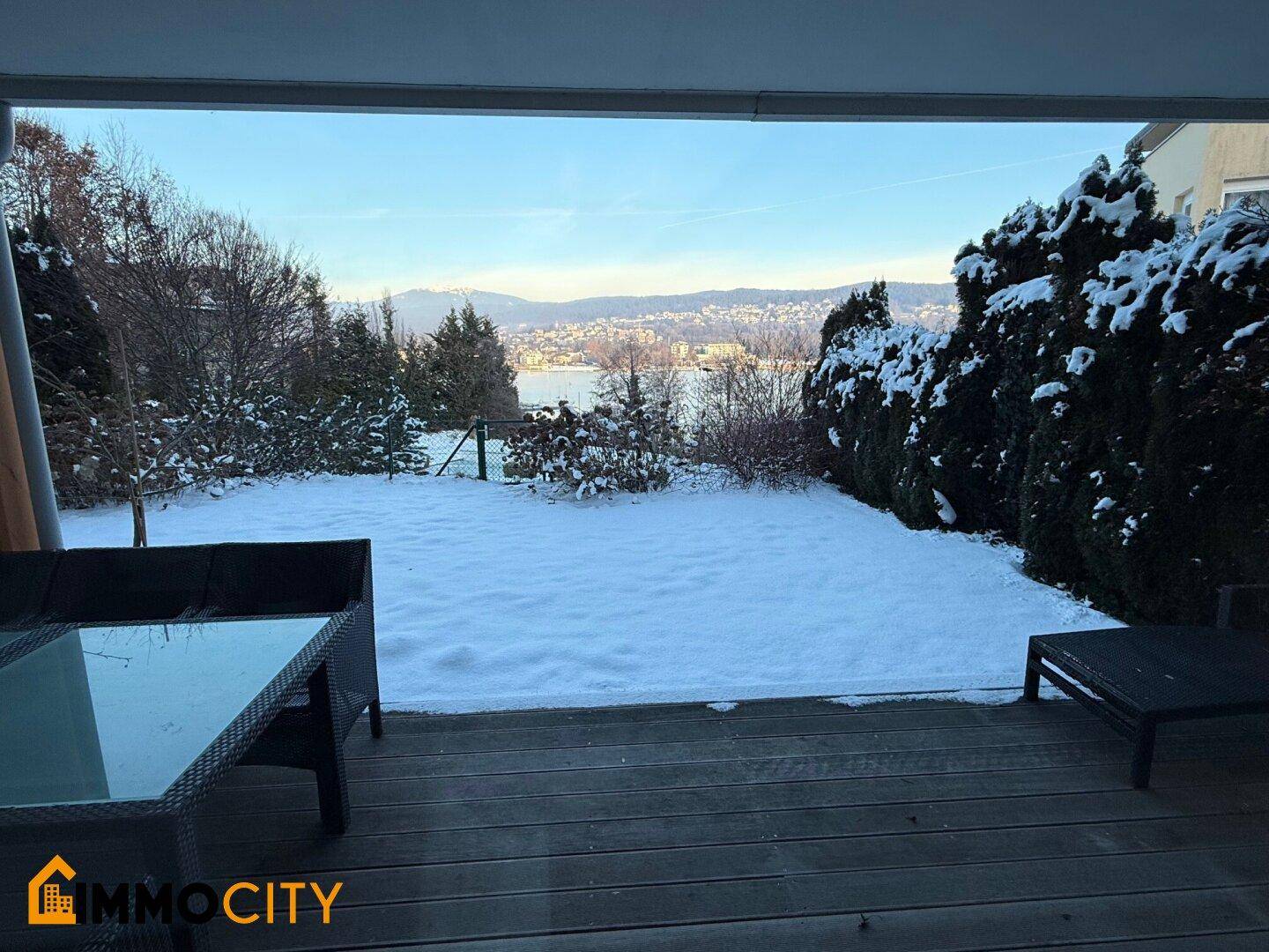 Gemütliche Holzterrasse mit Outdoor-Möbeln und weitem Blick über den schneebedeckten Garten zum See.