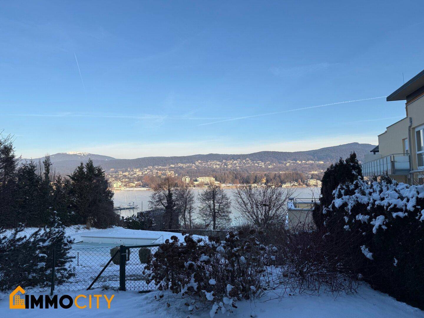 Winterlandschaft mit schneebedecktem Garten, See und Bergen, ergänzt durch einen Teil des modernen Gebäudes.