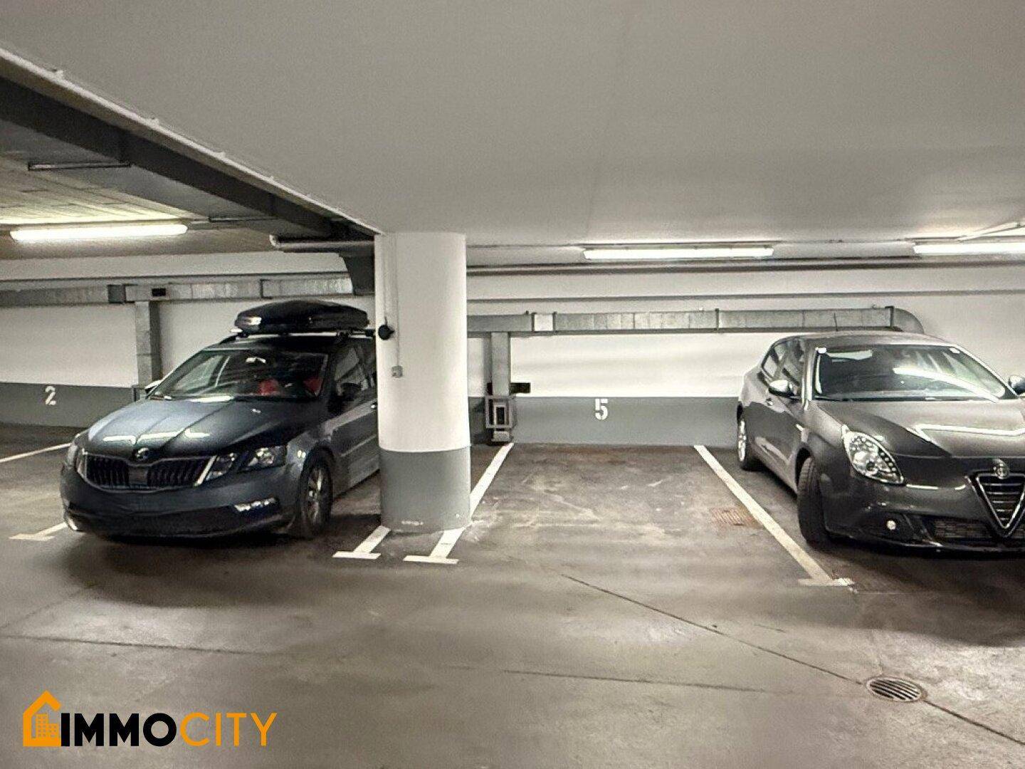 Tiefgarage mit zwei geparkten Autos, einem zentralen Pfeiler und markierten Stellplätzen.