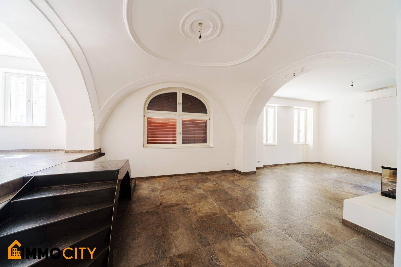 Helles Wohnzimmer mit großem Rundbogenfenster, Fliesenboden und Treppe zum erhöhten Bereich.