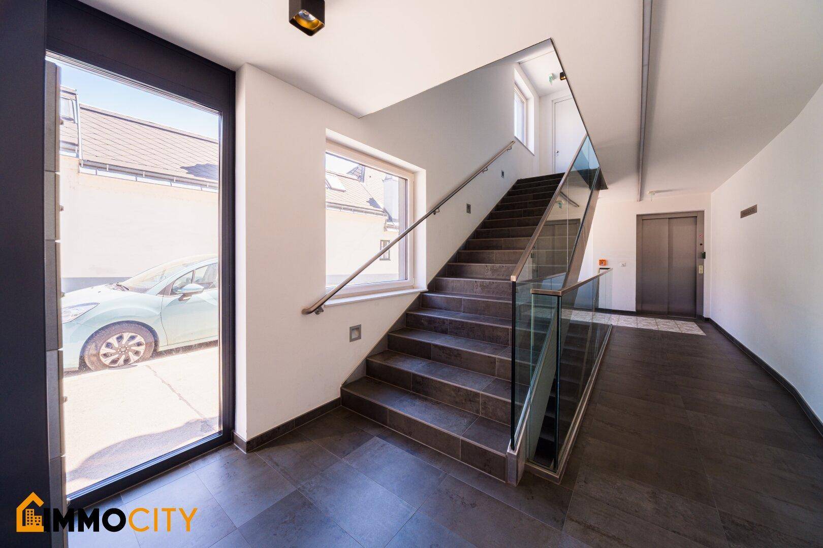Moderner Treppenaufgang mit Glasgeländer und Zugang zu einem Aufzug im Hintergrund.