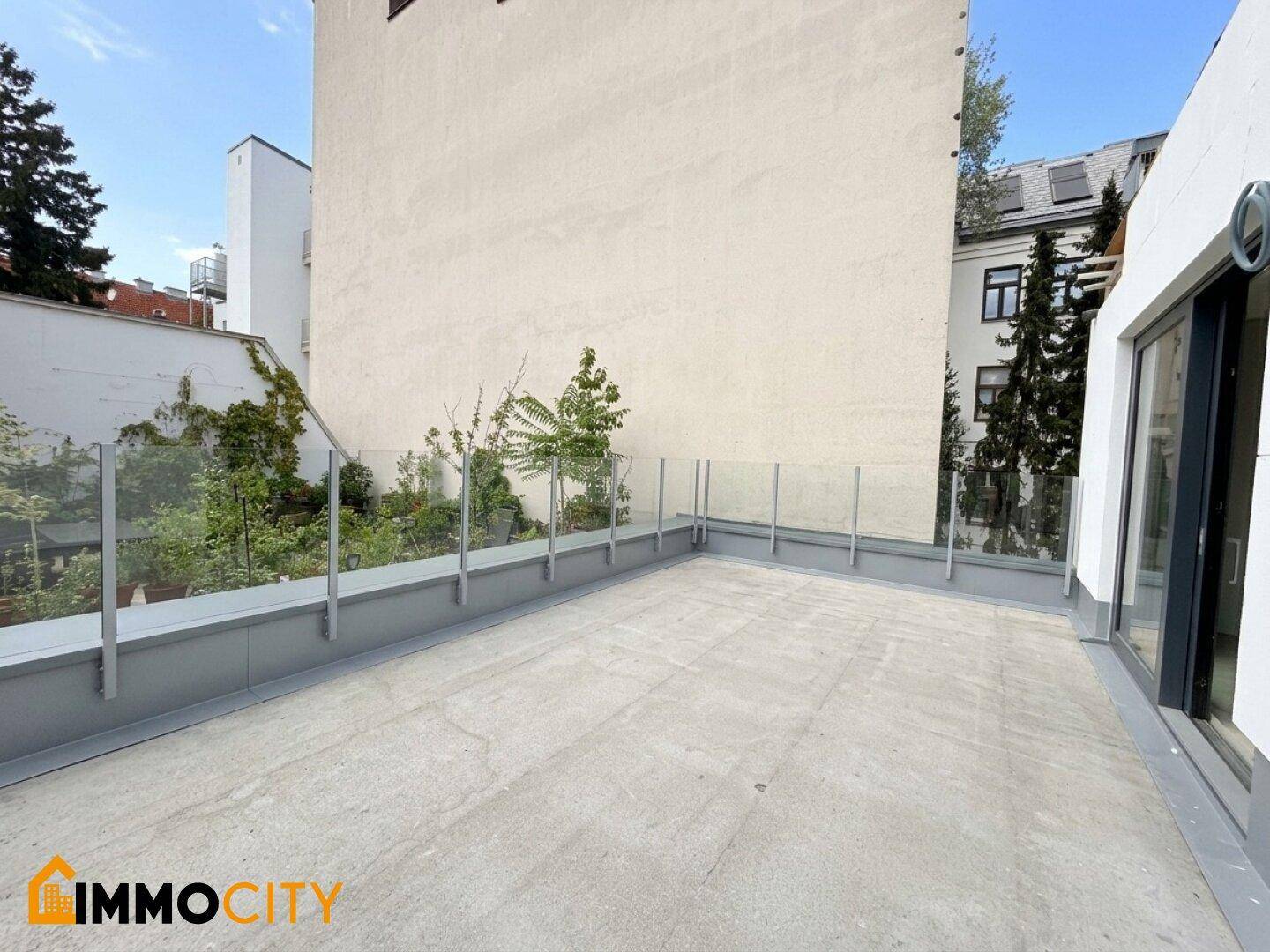 Weitläufige Terrasse mit modernem Glasgeländer und Blick auf die angrenzenden Gebäude und Bäume.