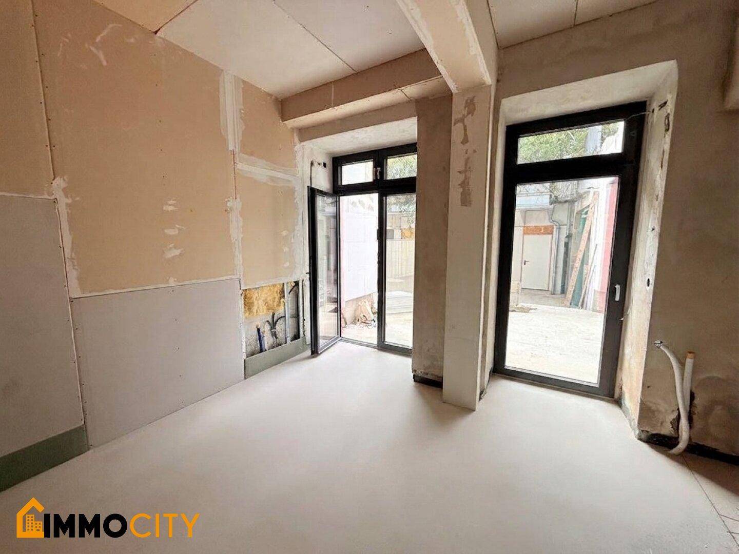 Unfertiger Raum mit großen Fenstertüren, die viel Tageslicht hereinlassen und einen Zugang nach draußen bieten.