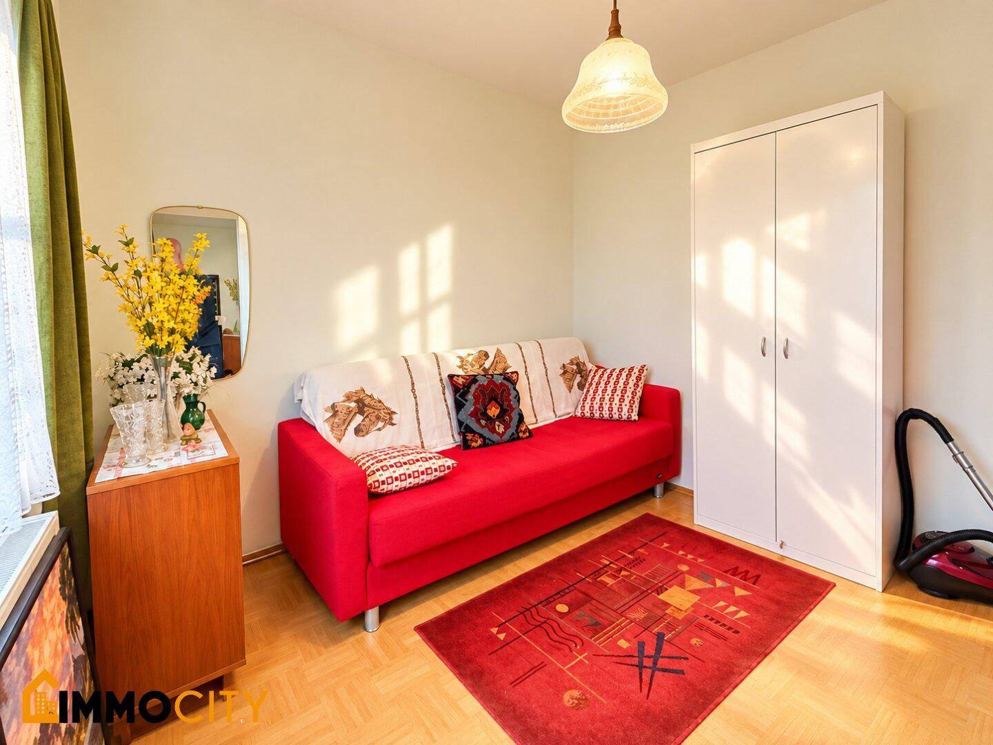 Helles Zimmer mit rotem Sofa, weißem Kleiderschrank und gemustertem Teppich.