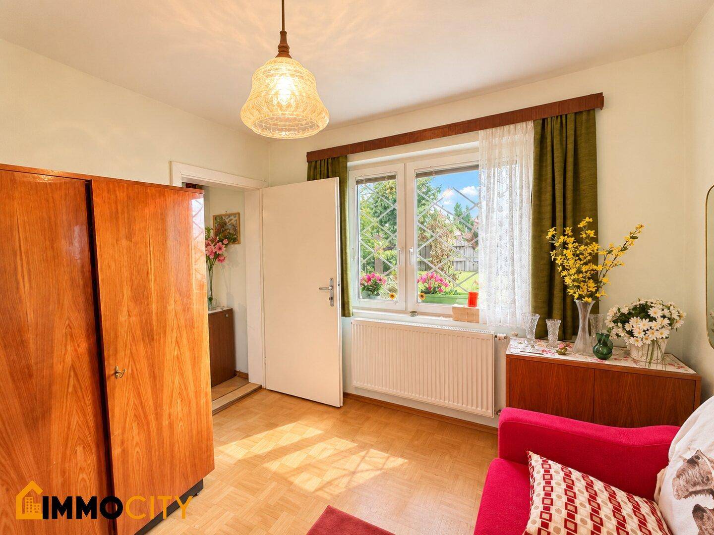 Helles Zimmer mit Holzschrank, rotem Sofa und Blick in den grünen Garten.