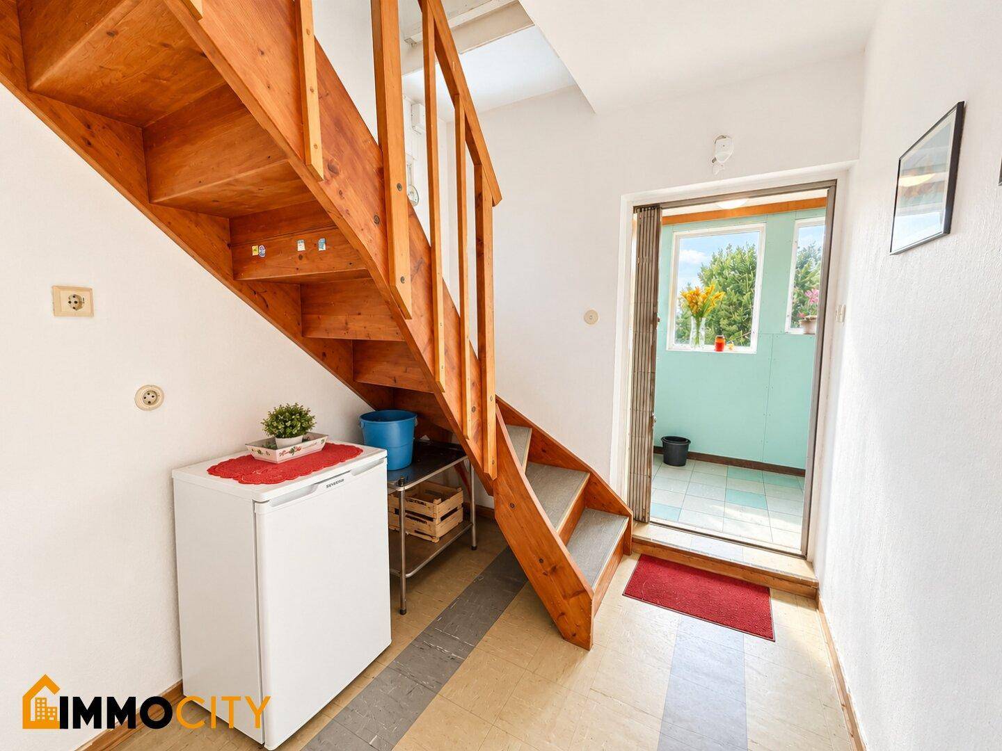 Heller Flur mit Holztreppe und Zugang zu einem verglasten Bereich mit Blick nach draußen.