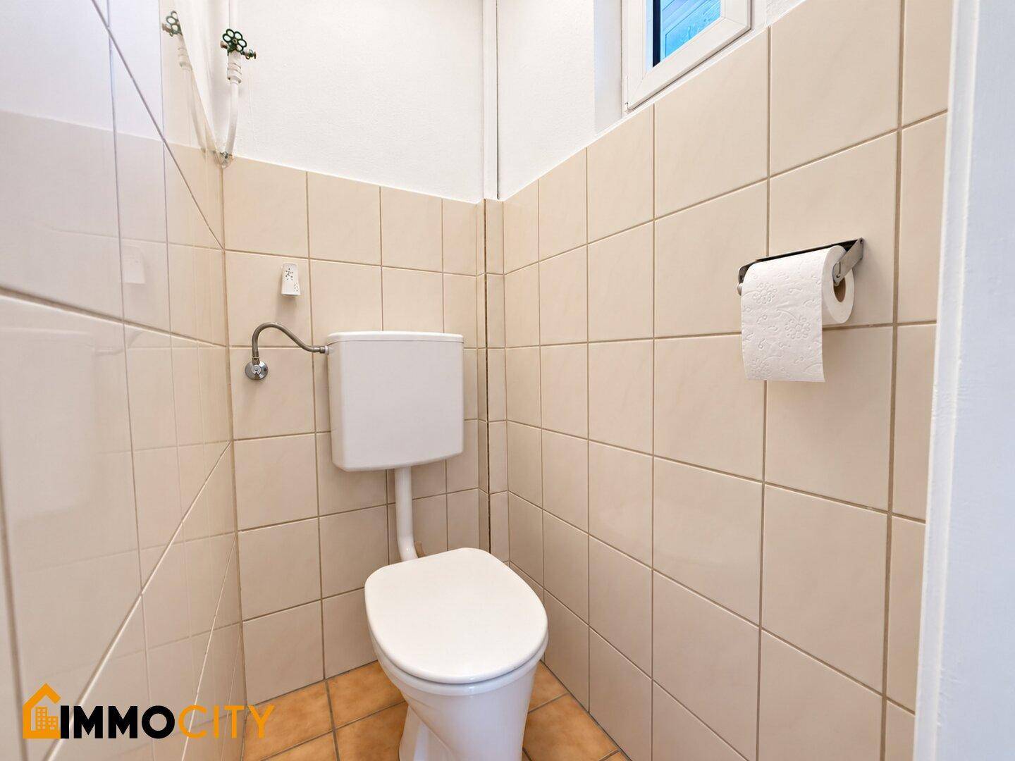 Kleines Gäste-WC mit beigefarbenen Fliesen und einem Fenster für natürliche Belüftung.