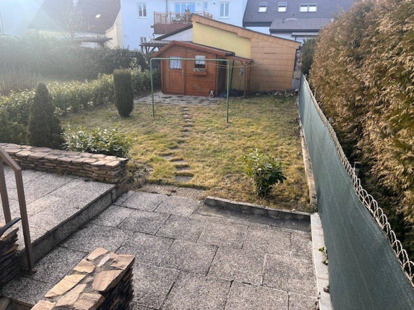 Gartenbereich mit gepflasterter Terrasse, Rasenfläche und Gartenhaus im Hintergrund.