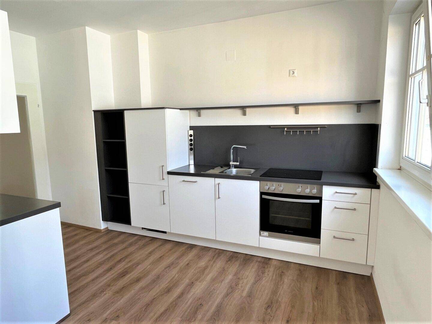 Moderne Einbauküche mit weißen Fronten, dunkler Arbeitsplatte und Laminatboden, hell durch Fenster.