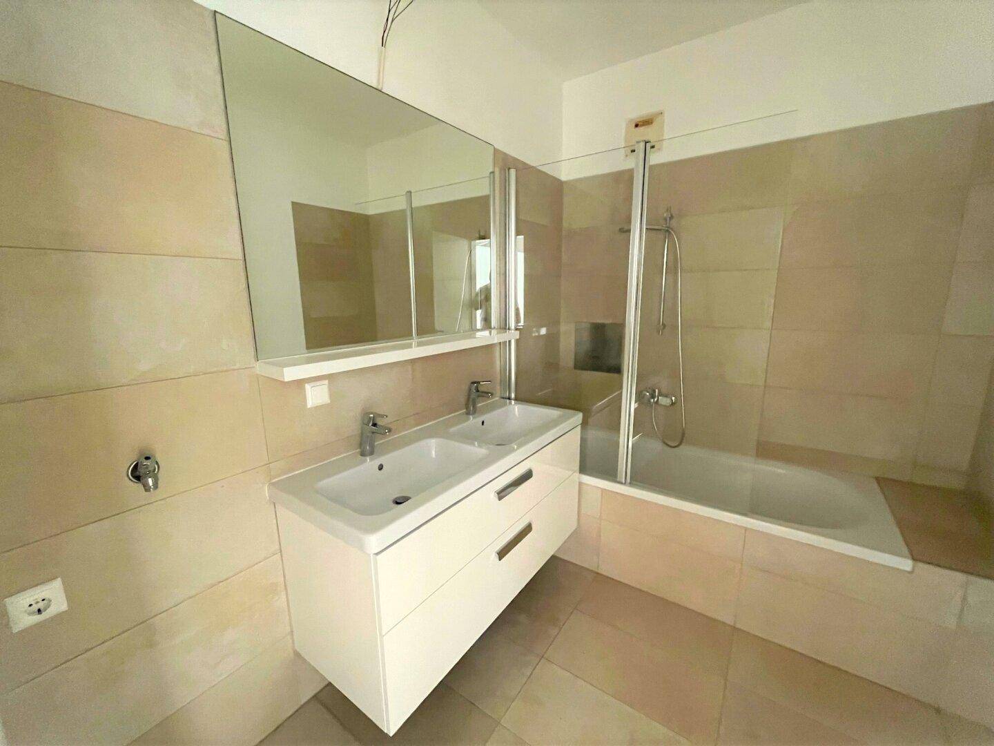 Helles Badezimmer mit beigefarbenen Fliesen, Doppelwaschbecken, Badewanne und Duschbereich.