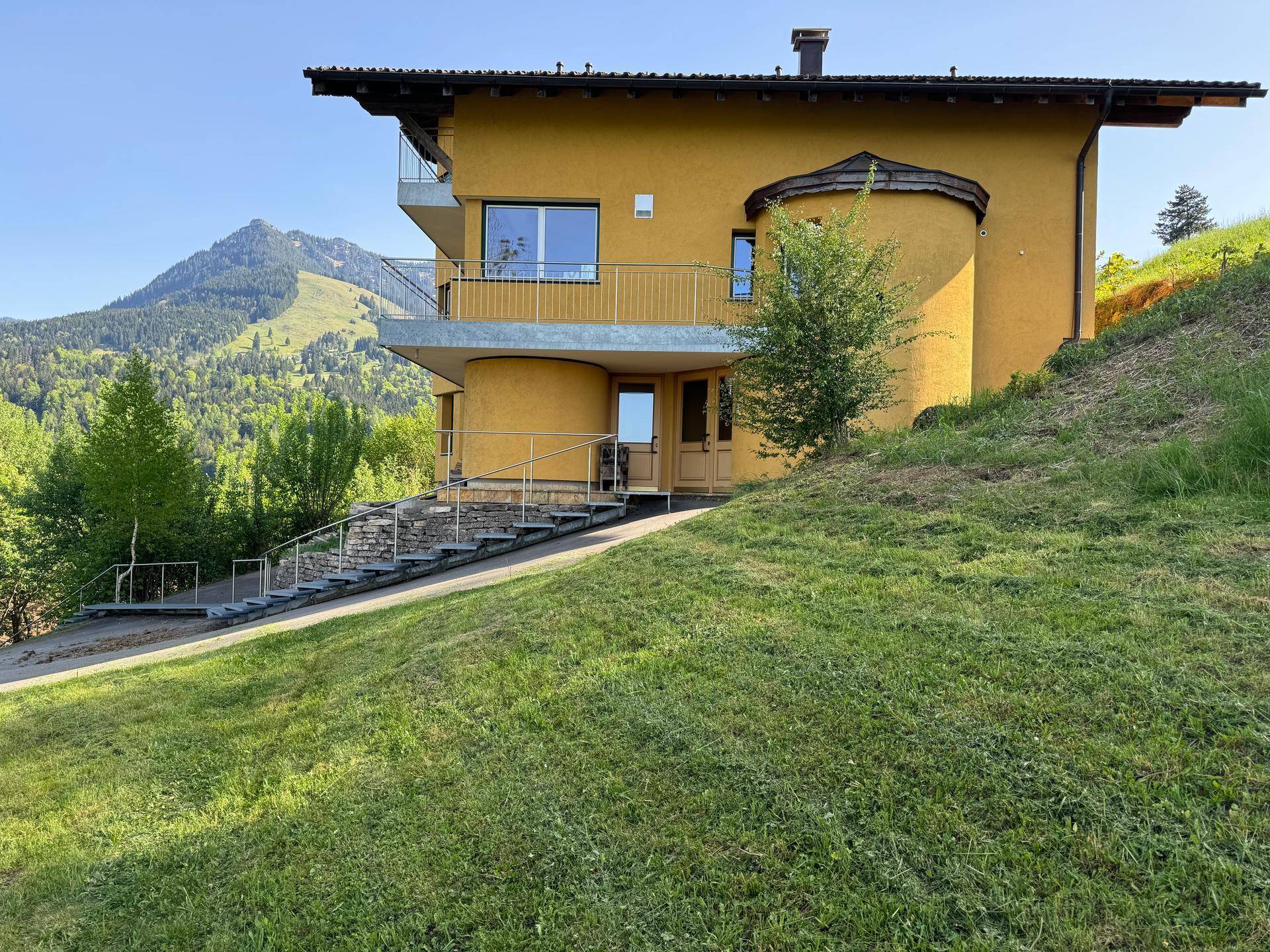 Gelbes Wohnhaus in Hanglage mit moderner Architektur und grüner Umgebung, umgeben von Bergen.