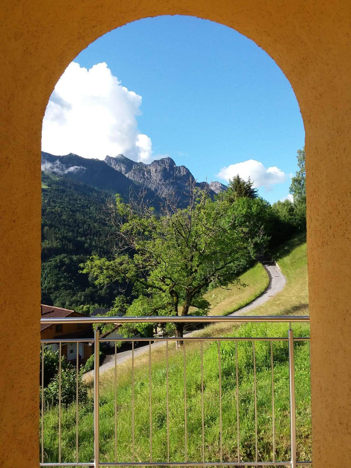 Malerischer Blick durch einen Bogen auf grüne Hügel und majestätische Berge unter blauem Himmel.