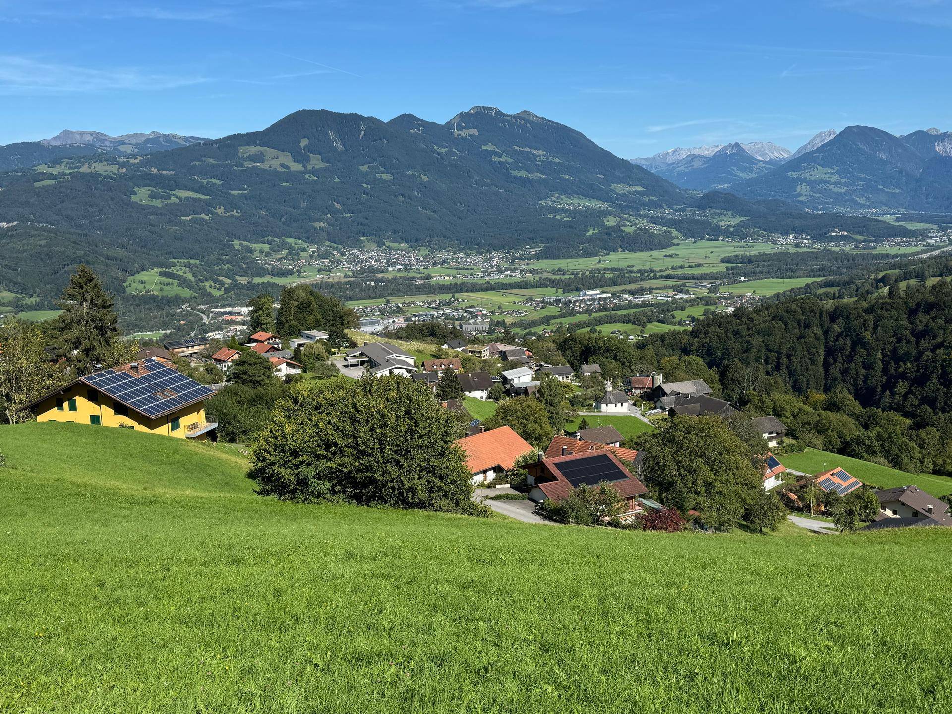 Weitläufiger Panoramablick über ein grünes Tal mit Siedlungen und majestätischen Bergen im Hintergrund.