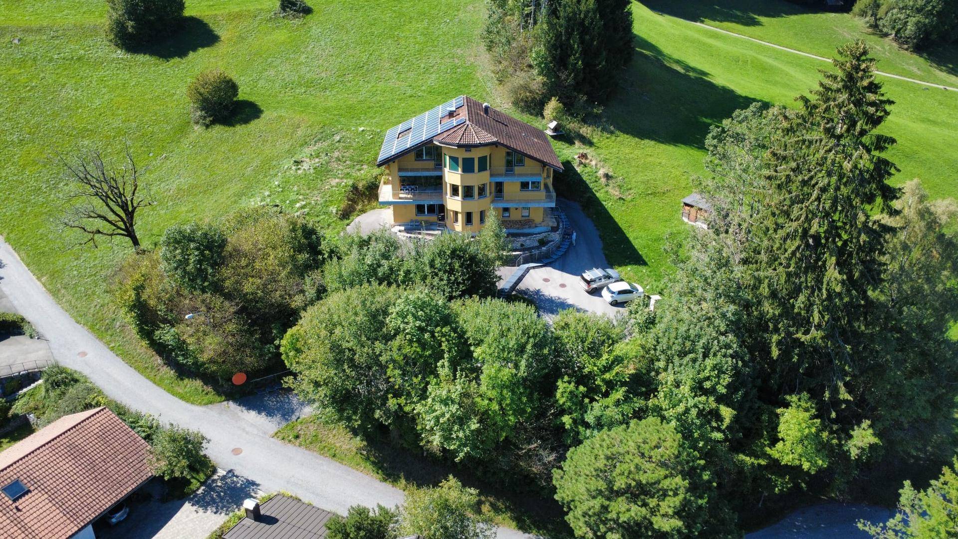 Luftaufnahme eines gelben Hauses mit Solaranlage auf dem Dach, eingebettet in grüne Hügel und Bäume.