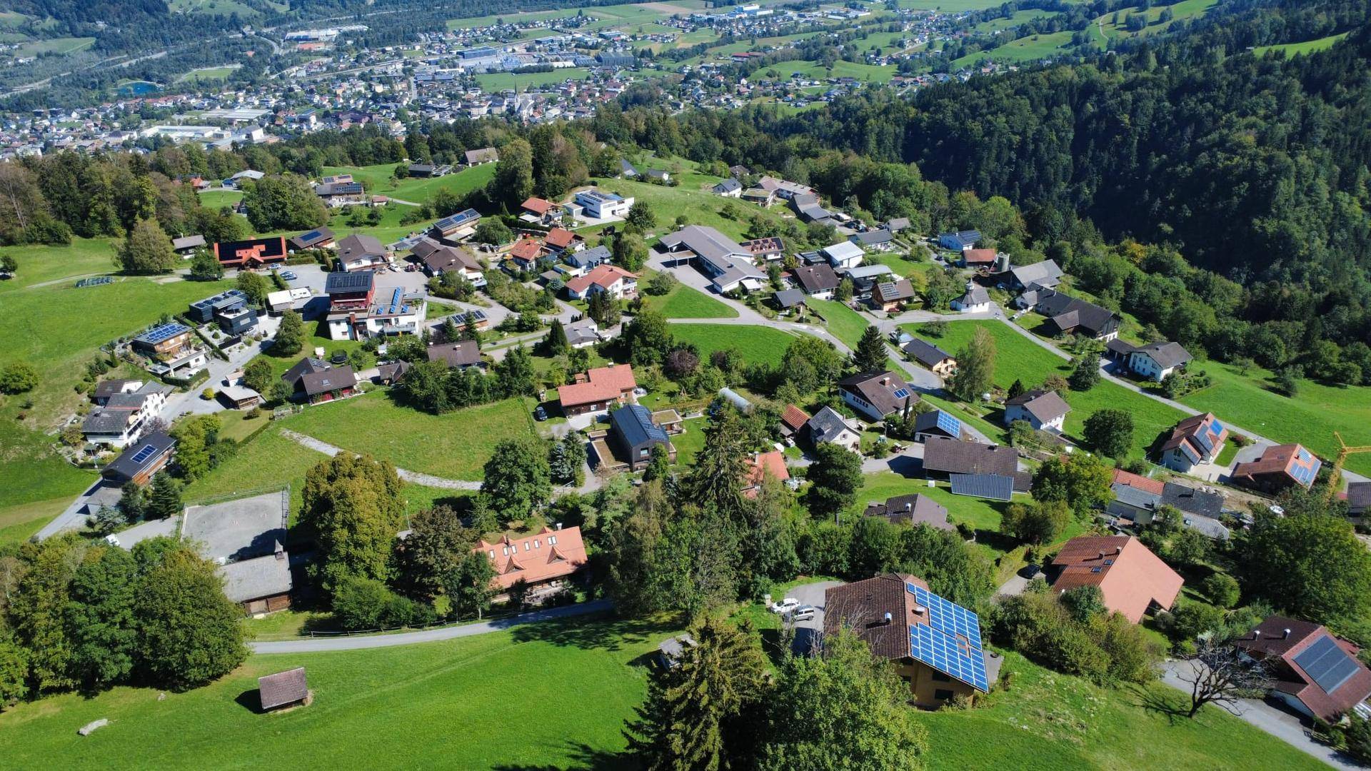 Weitläufige Luftaufnahme einer ländlichen Siedlung mit vielen Häusern und grüner Hügellandschaft.
