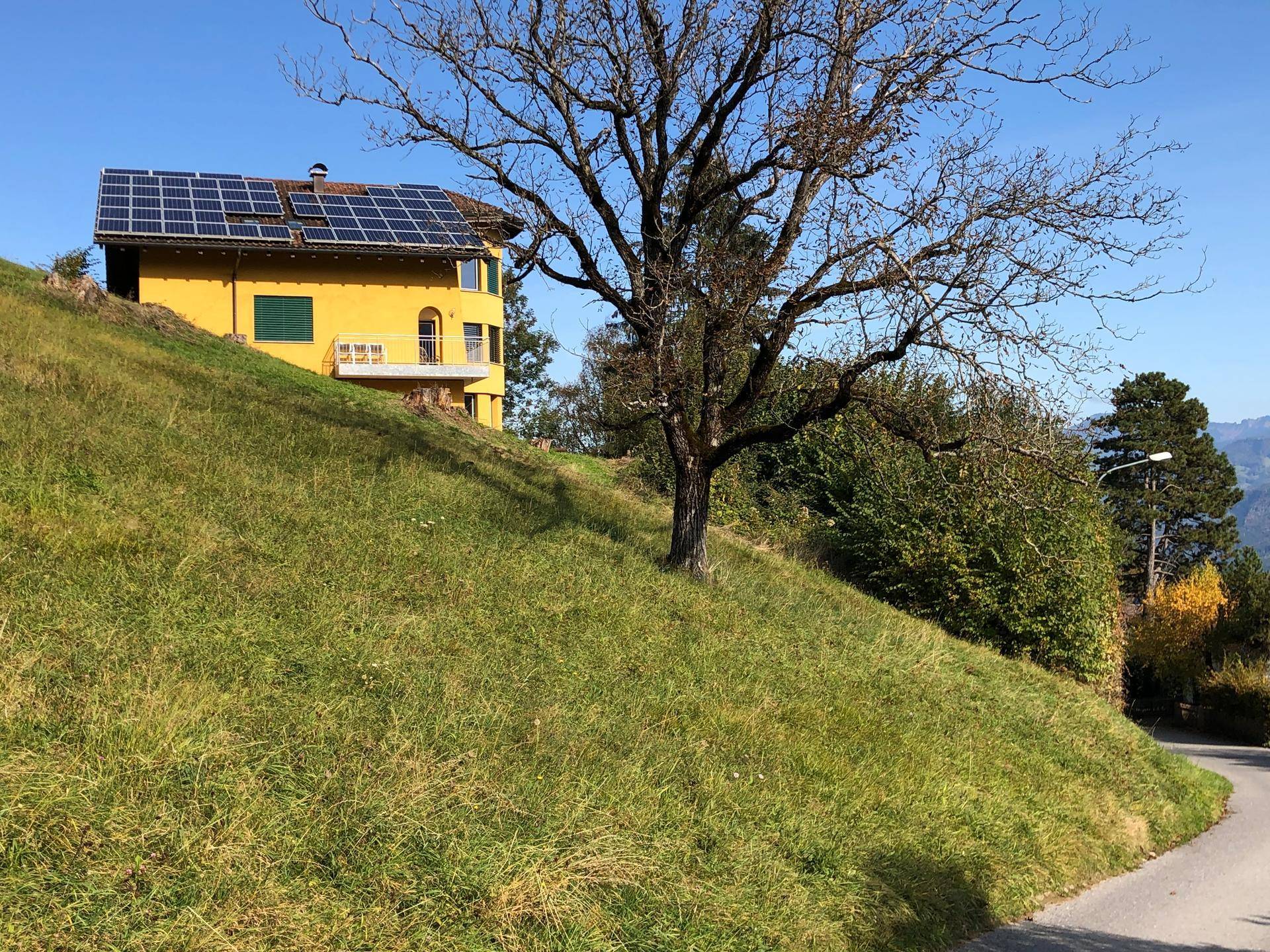 Gelbes Wohnhaus in Hanglage mit Photovoltaikanlage auf dem Dach, umgeben von grüner Natur.