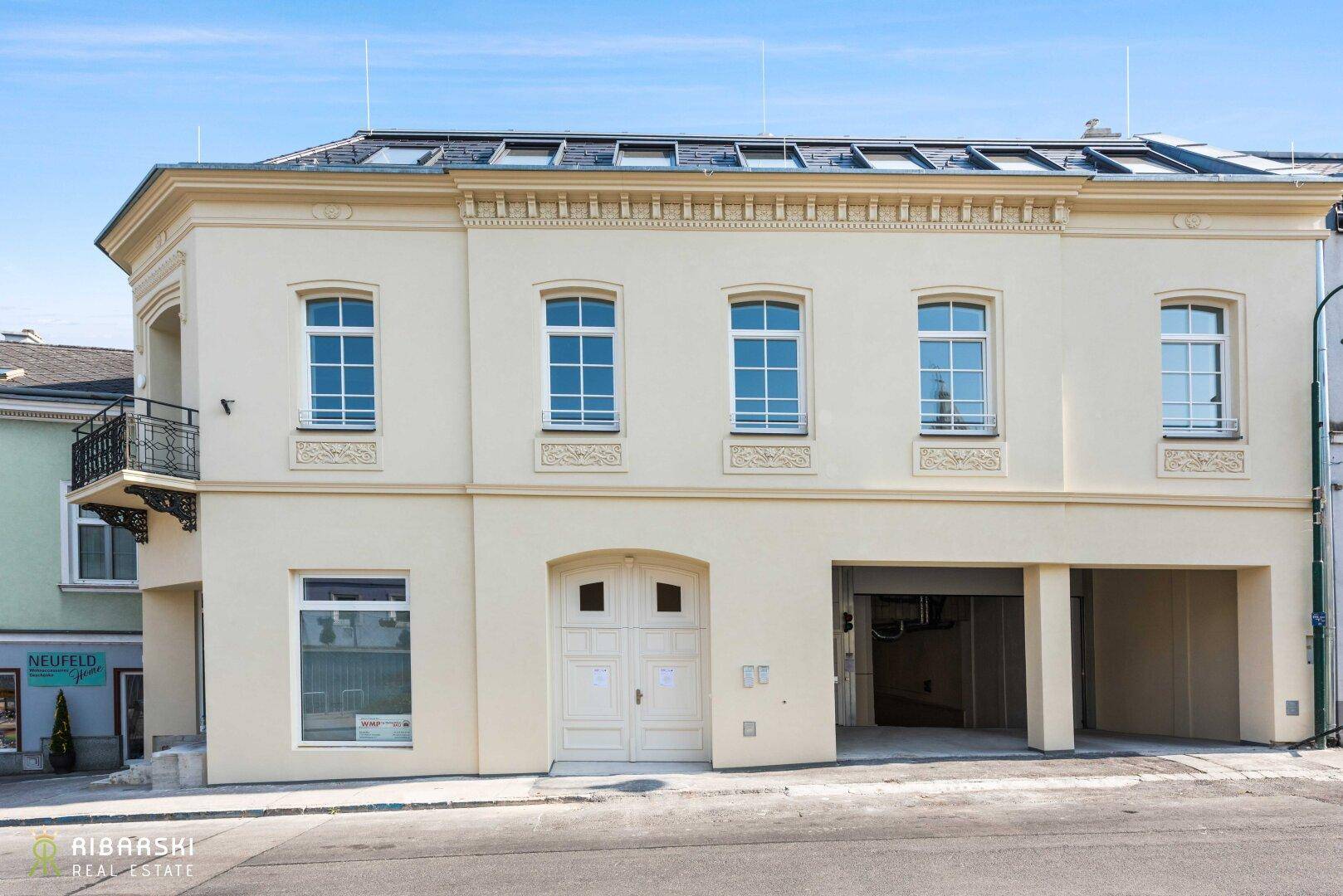 Gebäudeansicht von der Straße mit heller Fassade, Bogenfenstern und Garageneinfahrt.