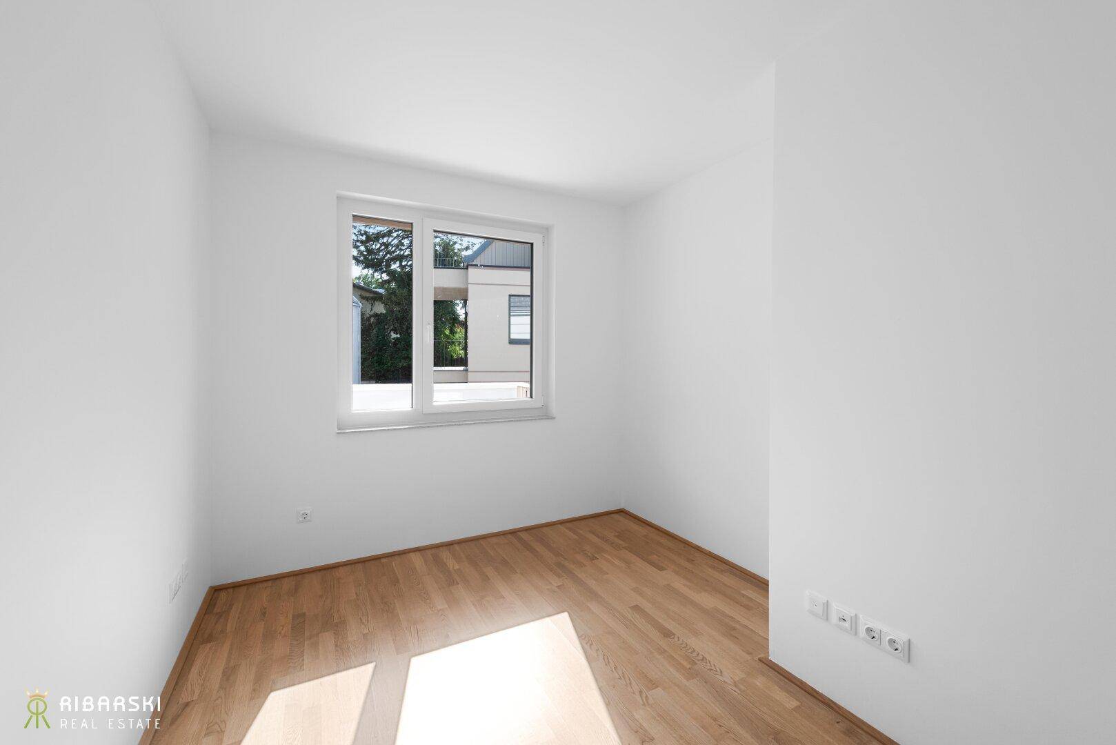 Helles Zimmer mit Parkettboden und Fenster, das einen Blick ins Grüne bietet.