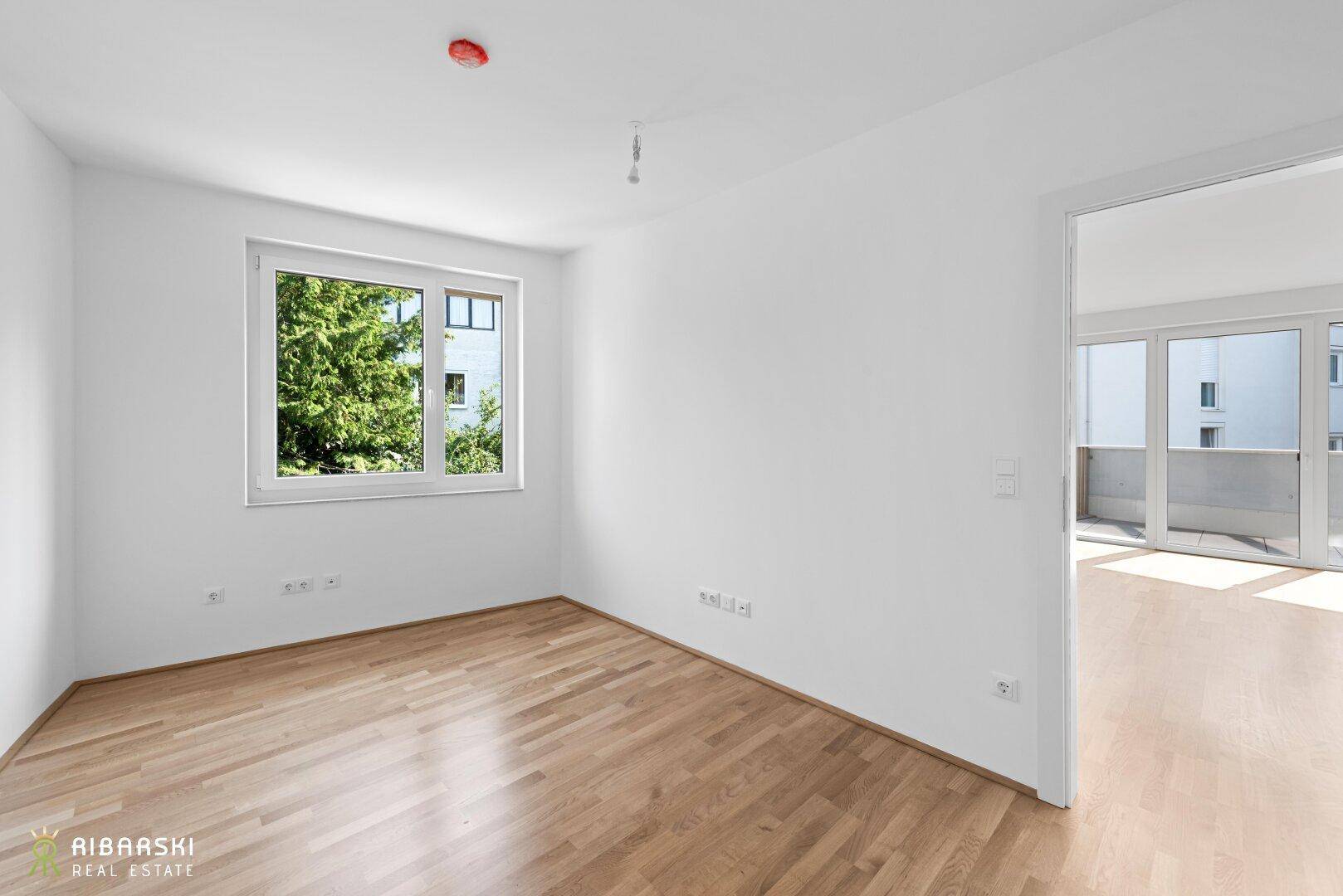 Lichtdurchflutetes Zimmer mit Parkettboden und Zugang zu einem weiteren Raum mit Balkon.
