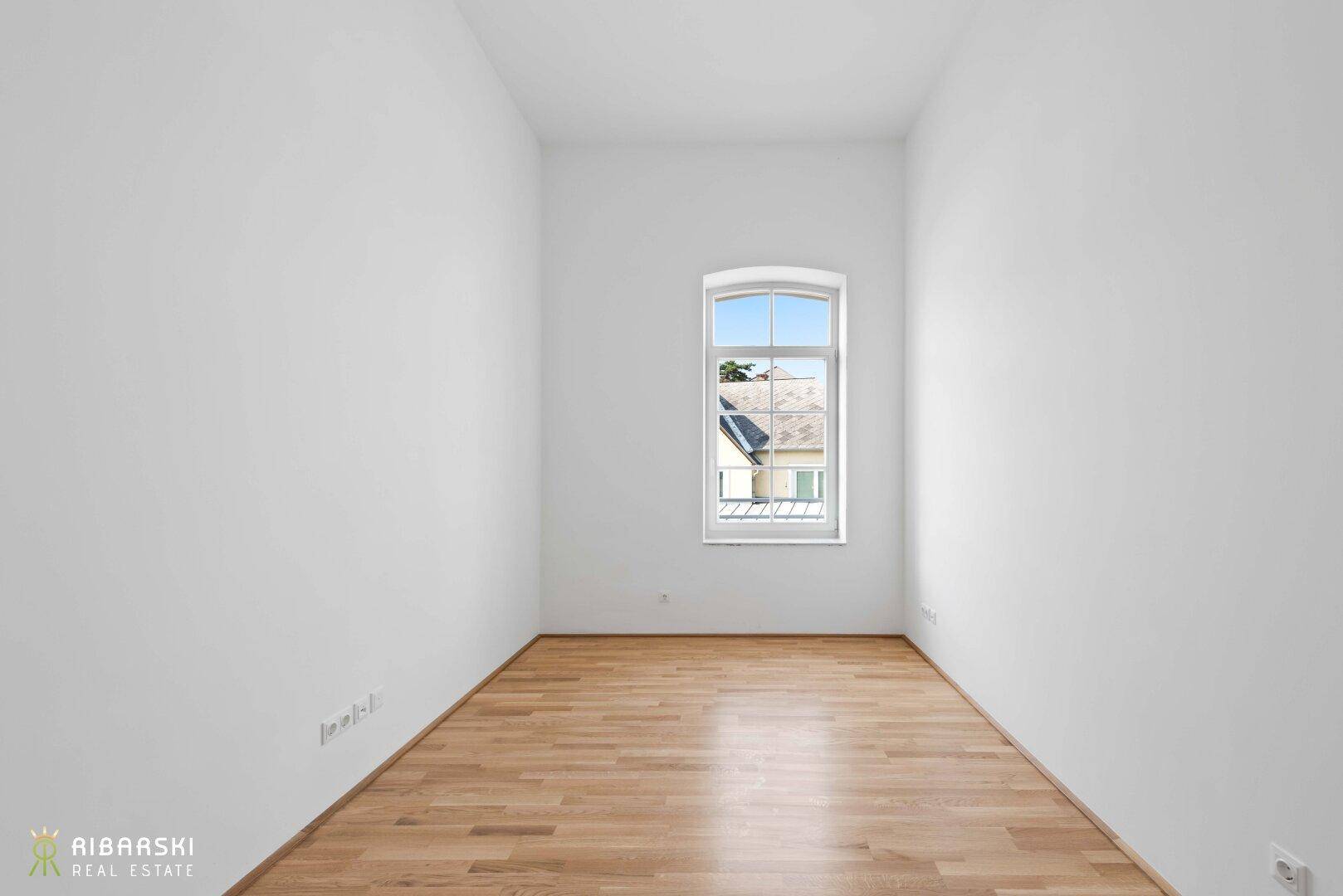 Leeres Zimmer mit hellen Wänden, Parkettboden und einem Fenster mit Blick ins Grüne.