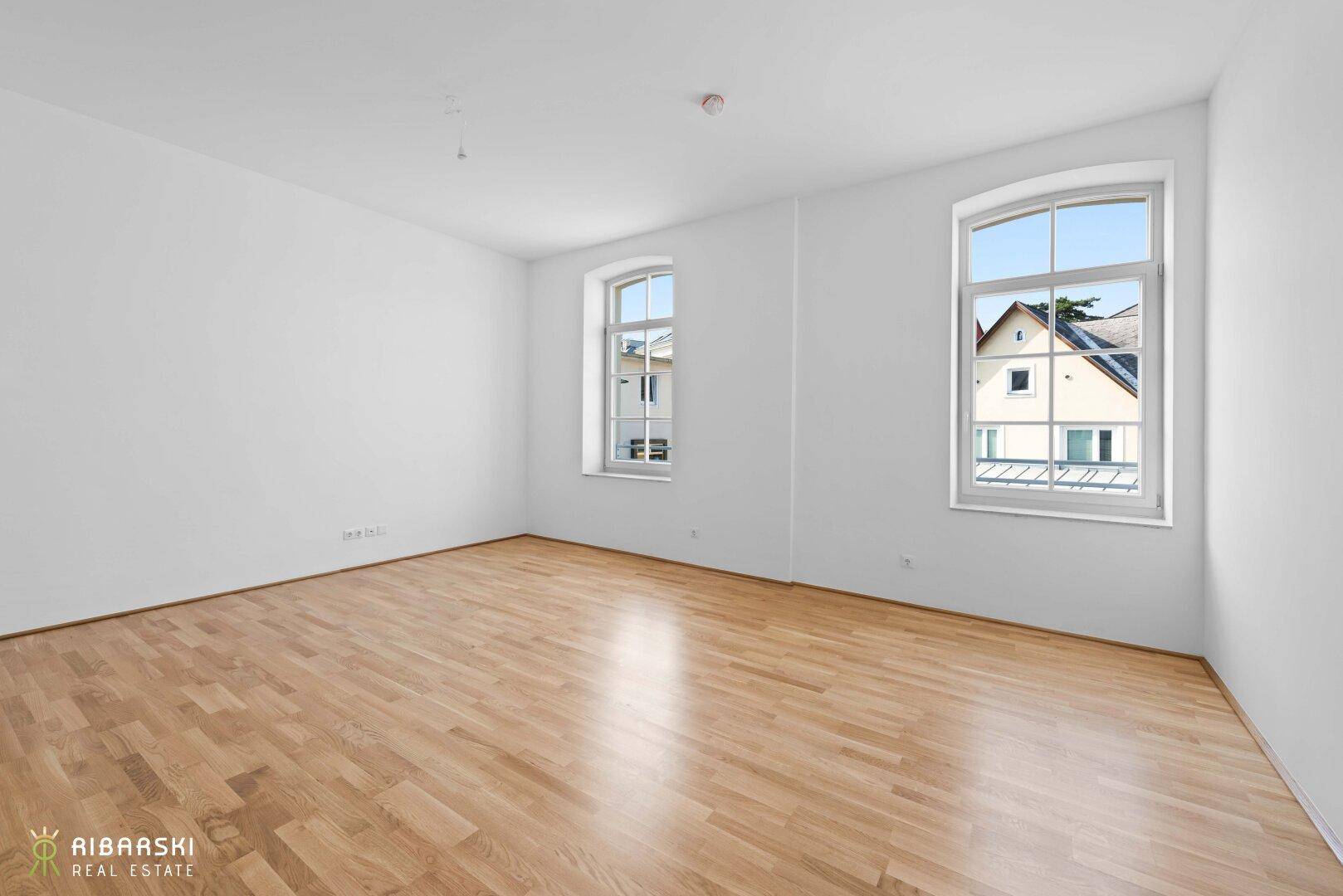 Geräumiges Zimmer mit Parkettboden und zwei Fenstern, die viel Tageslicht hereinlassen.