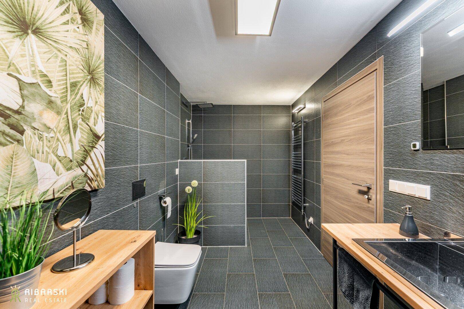Modernes Badezimmer mit dunklen Fliesen, begehbarer Dusche und wandhängendem WC.