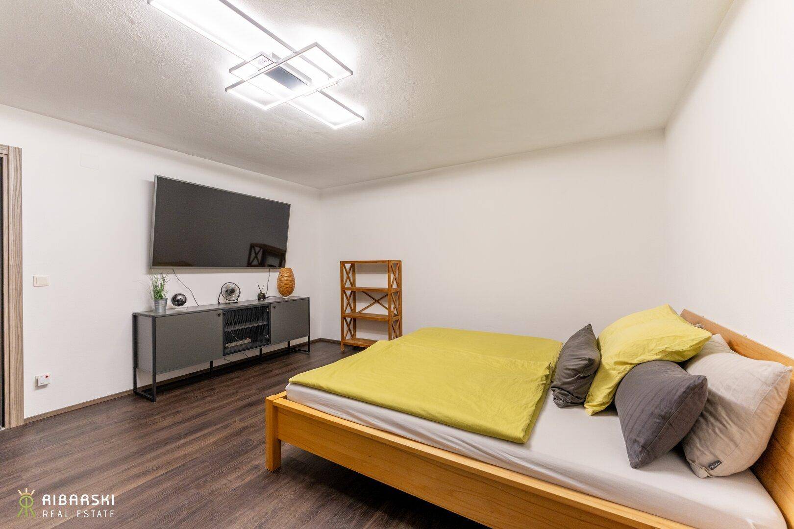 Einfaches Schlafzimmer mit Holzbett, Holzboden und großem Wandfernseher.