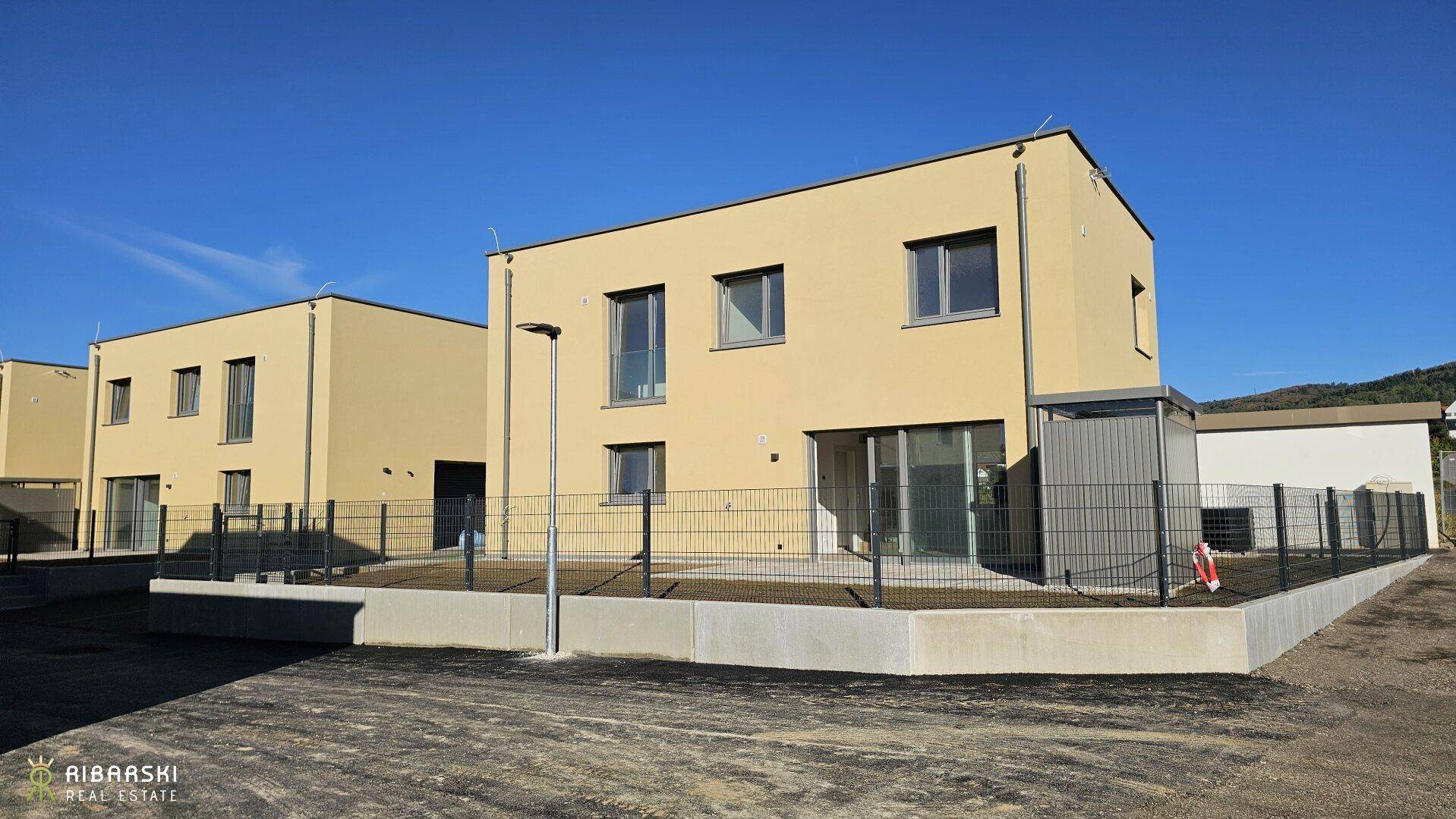 Neubau-Doppelhaushälfte mit heller Fassade, modernem Design und kleinem Gartenbereich unter blauem Himmel.