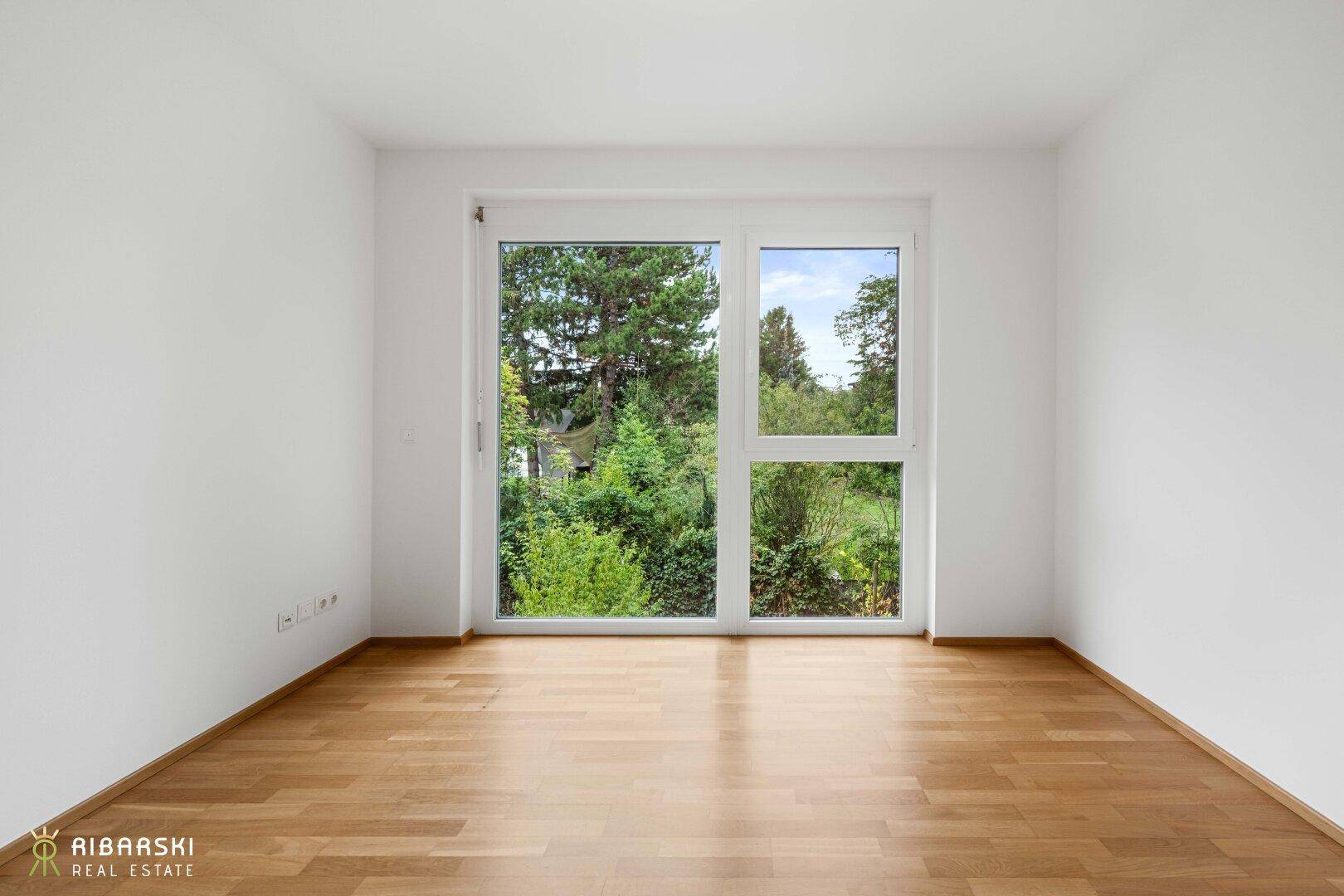 Helles Zimmer mit Parkettboden und großem Fenster mit Blick ins Grüne.