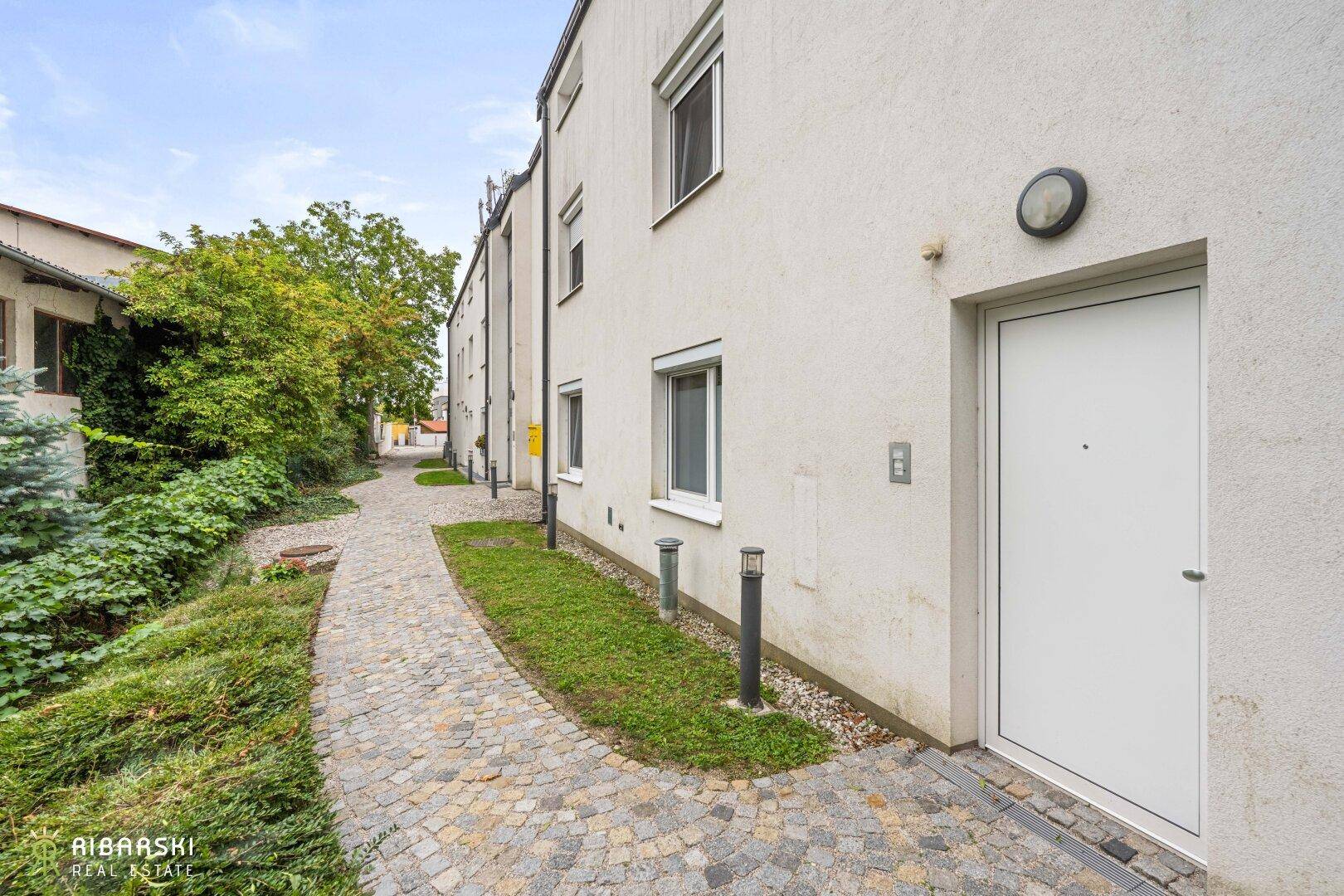 Gepflasterter Weg entlang der Gebäudeseite mit Eingangstür und Grünflächen.