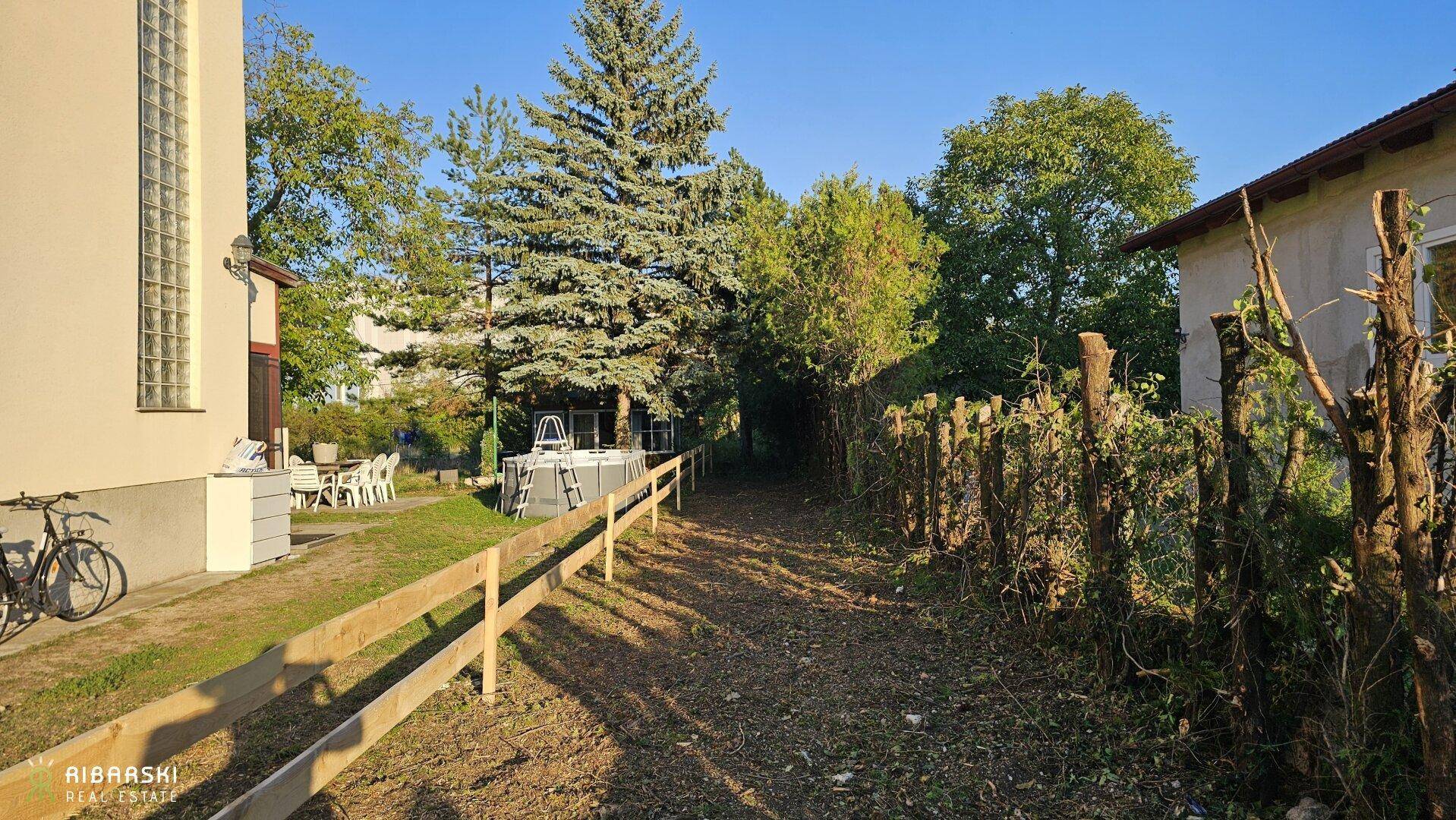 Gartenweg mit Holzzaun, vorbei an einem Haus und einem aufgestellten Swimmingpool.