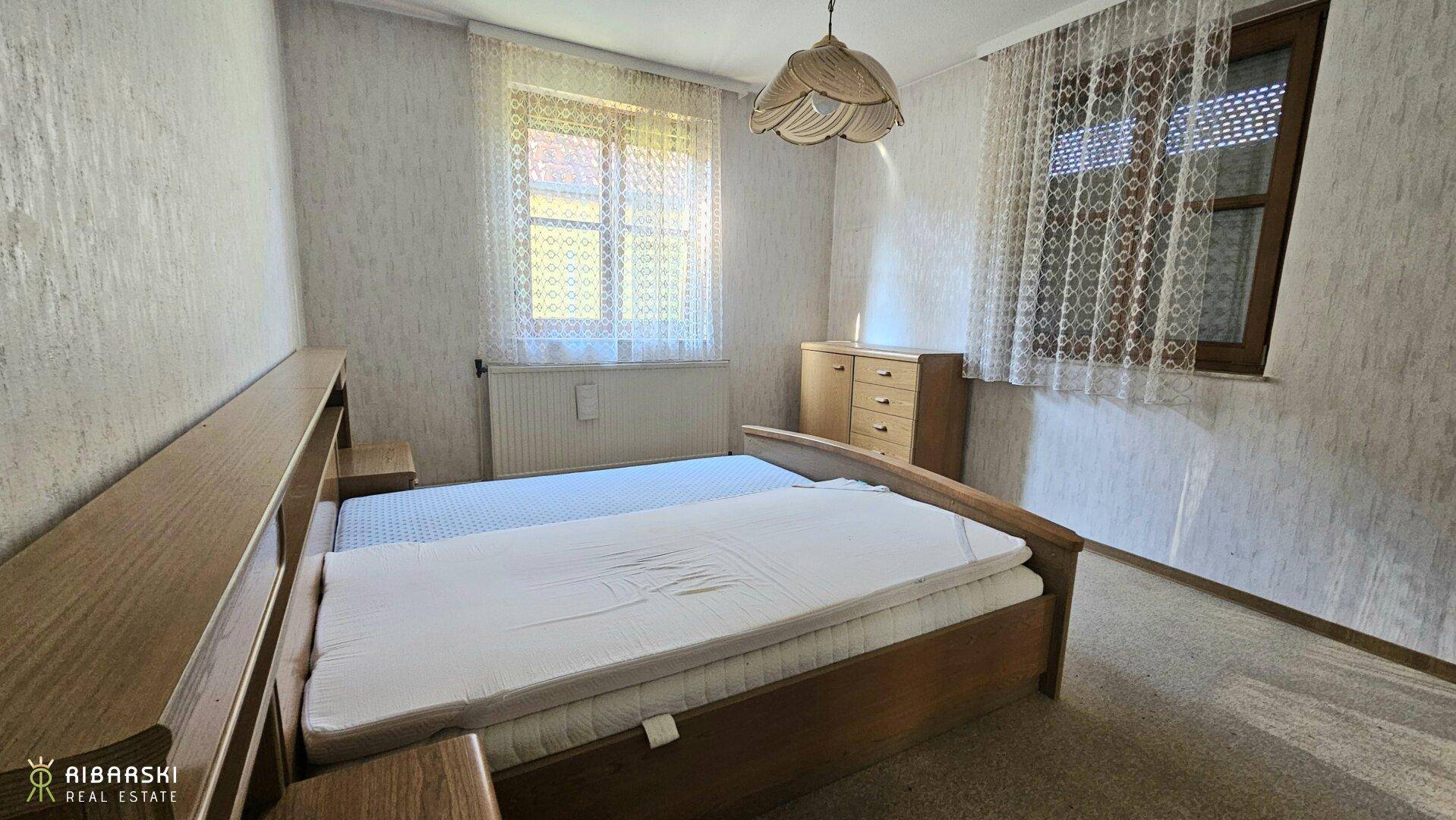 Schlafzimmer mit Doppelbett und Holzmöbeln, Teppichboden und zwei Fenstern für natürliches Licht.