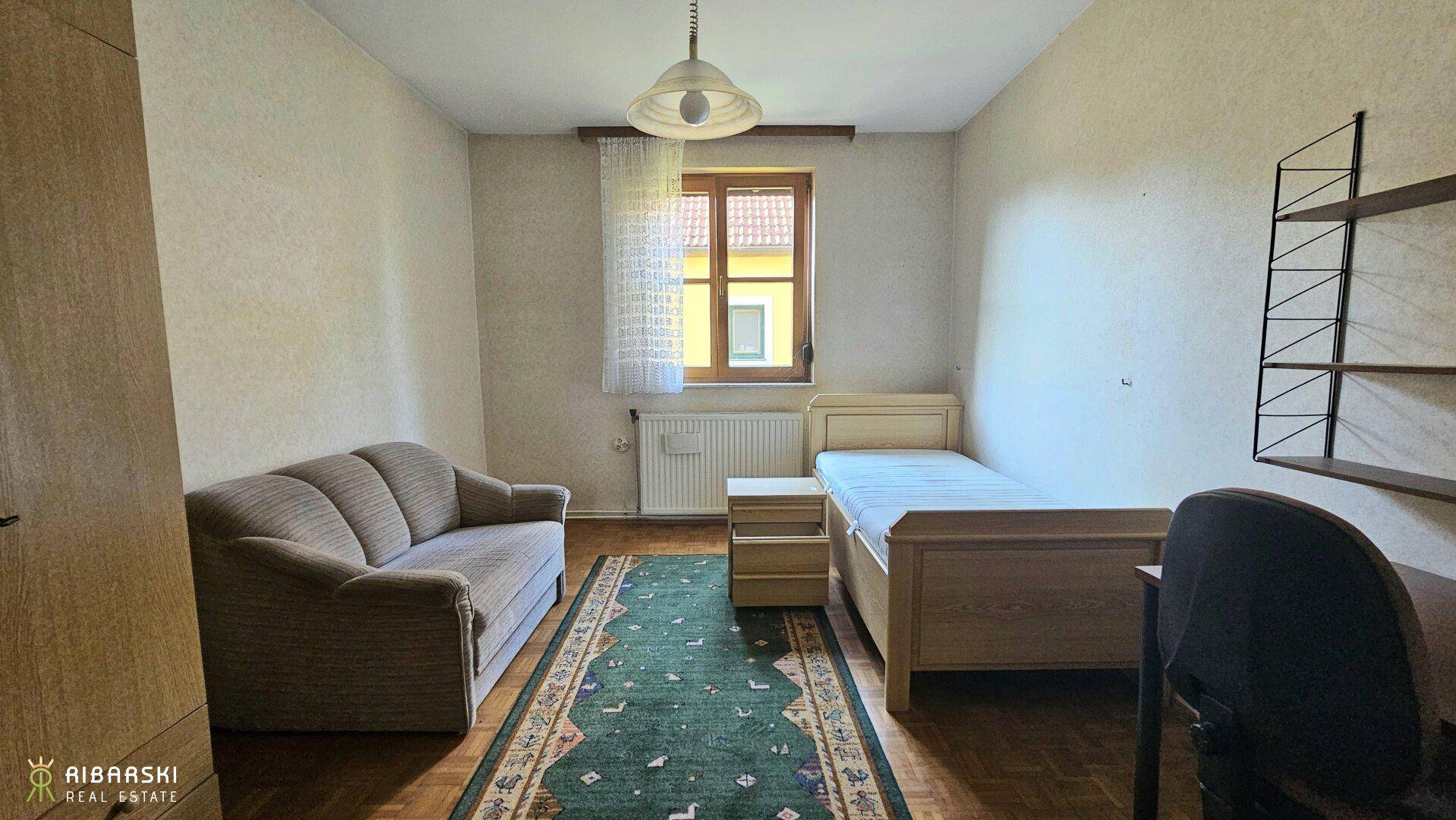 Schlafzimmer mit Einzelbett, Sofa und Schreibtisch, ausgestattet mit Holzmöbeln und Teppich.