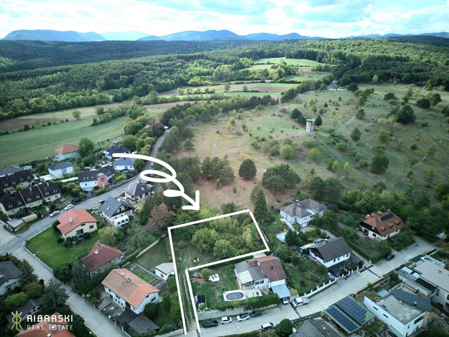 Luftaufnahme eines umrandeten Grundstücks mit umliegenden Häusern und weitreichender grüner Landschaft im Hintergrund.