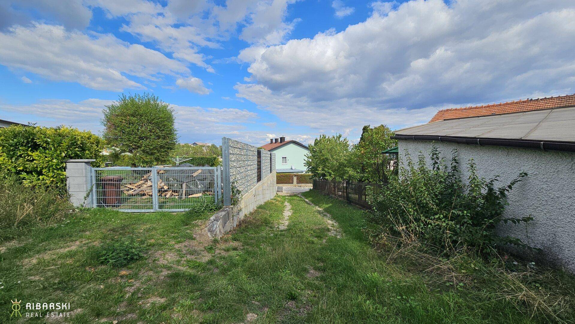 Grüner Weg führt zu einem Tor, umgeben von Hecken und einem Haus auf der rechten Seite.
